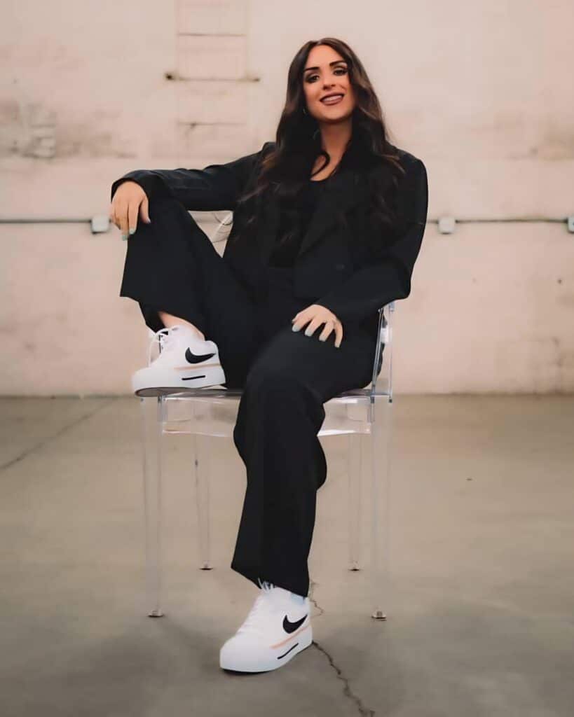 A woman sitting confidently on a transparent chair in a spacious room. She is wearing a black outfit with loose pants and a top, paired with white Nike sneakers. The background features a simple, industrial setting.