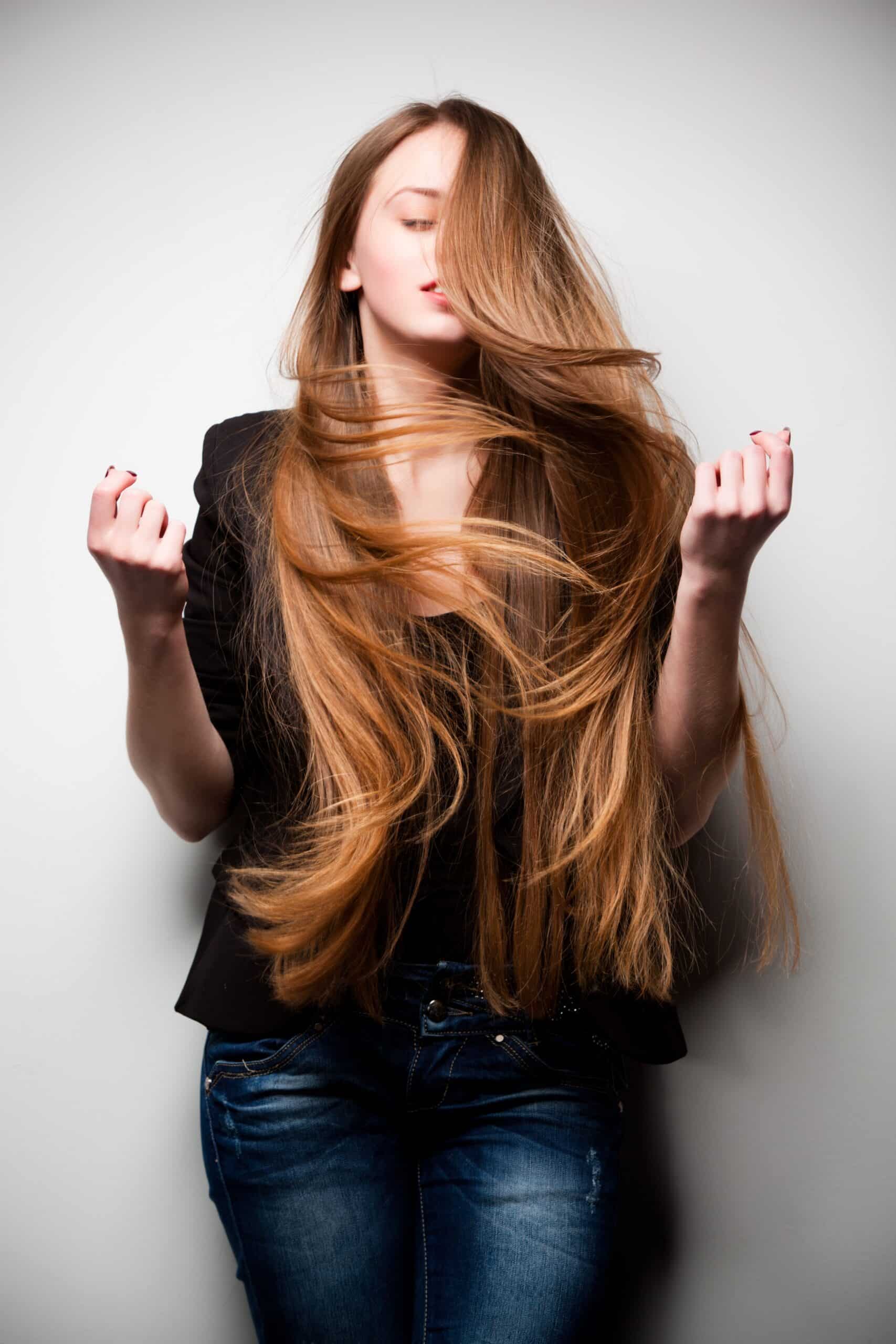 A woman with long, flowing hair stands against a plain backdrop, wearing a black jacket and denim jeans. She is holding her hair up with both hands, creating a sense of movement in the image.
