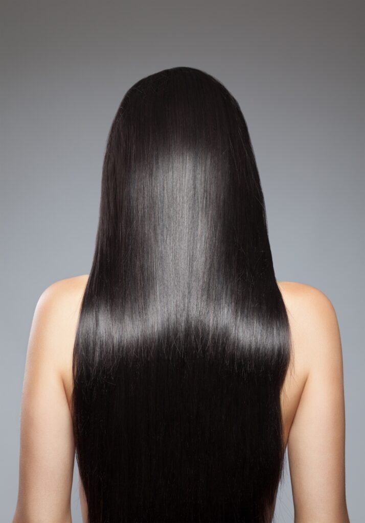 A close-up view of long, straight black hair from behind, with a smooth and shiny texture, set against a neutral gray background.