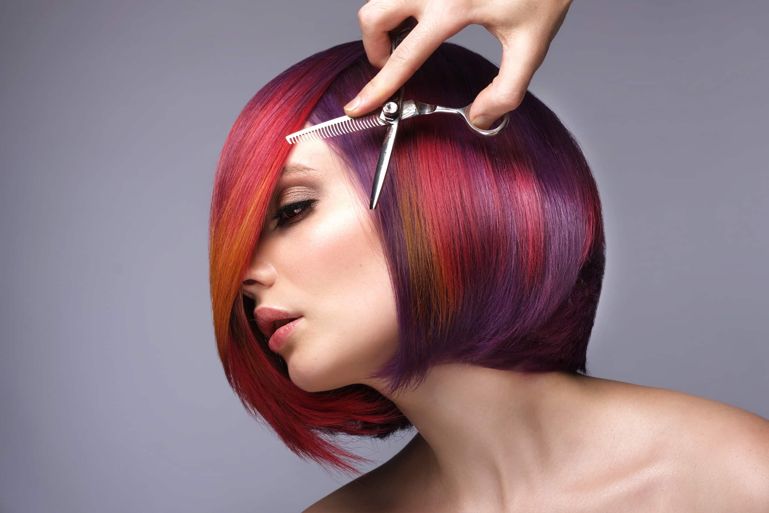 A close-up of a woman with a colorful bob haircut featuring shades of red, purple, and orange. A hand is holding scissors near her hair, indicating a hairstyle transformation. The background is a neutral gray.