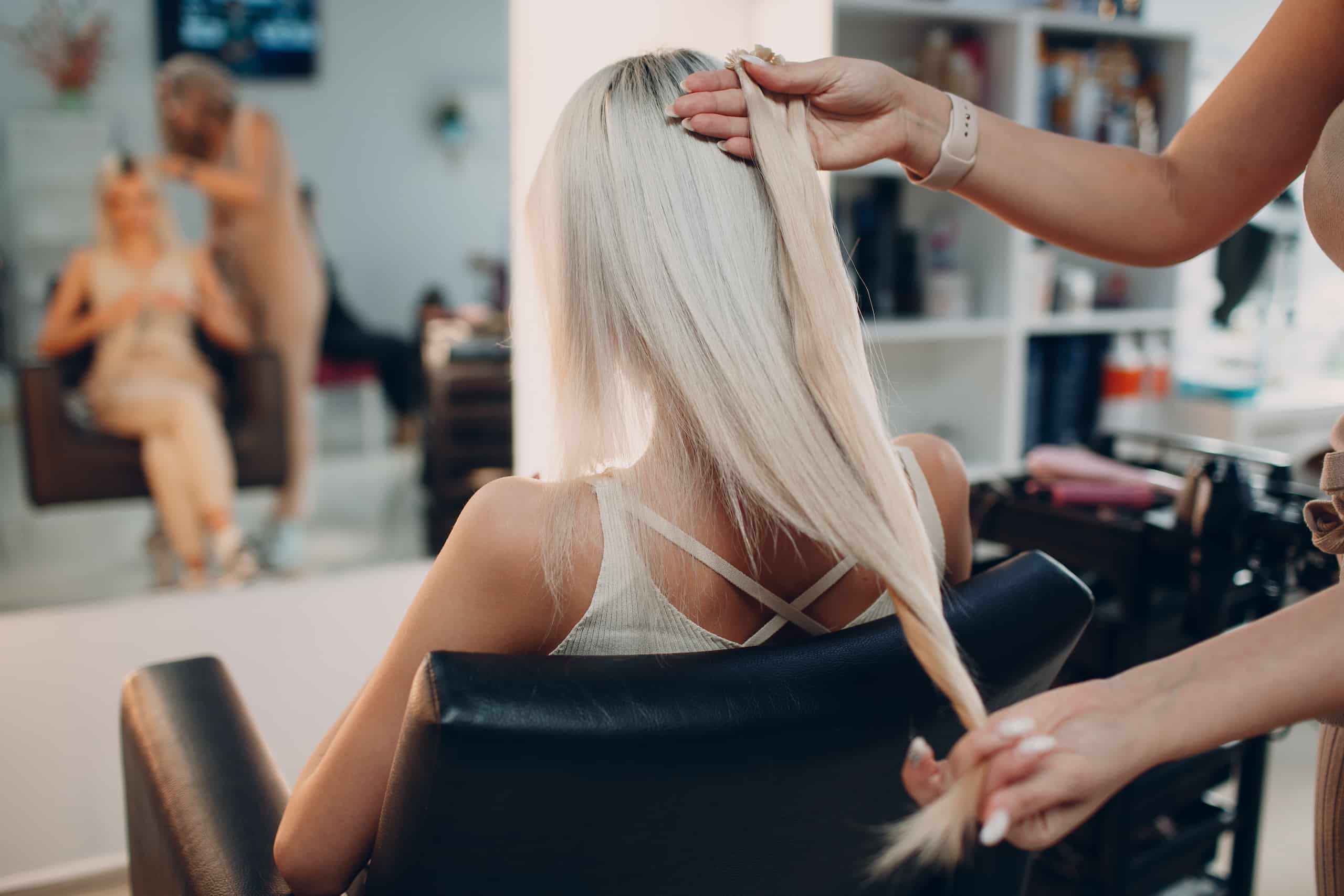 A hair stylist pulling a long strand of straight, light blonde hair from a client seated in a salon chair. The client is facing away, and the reflection in the mirror shows another stylist working nearby.
