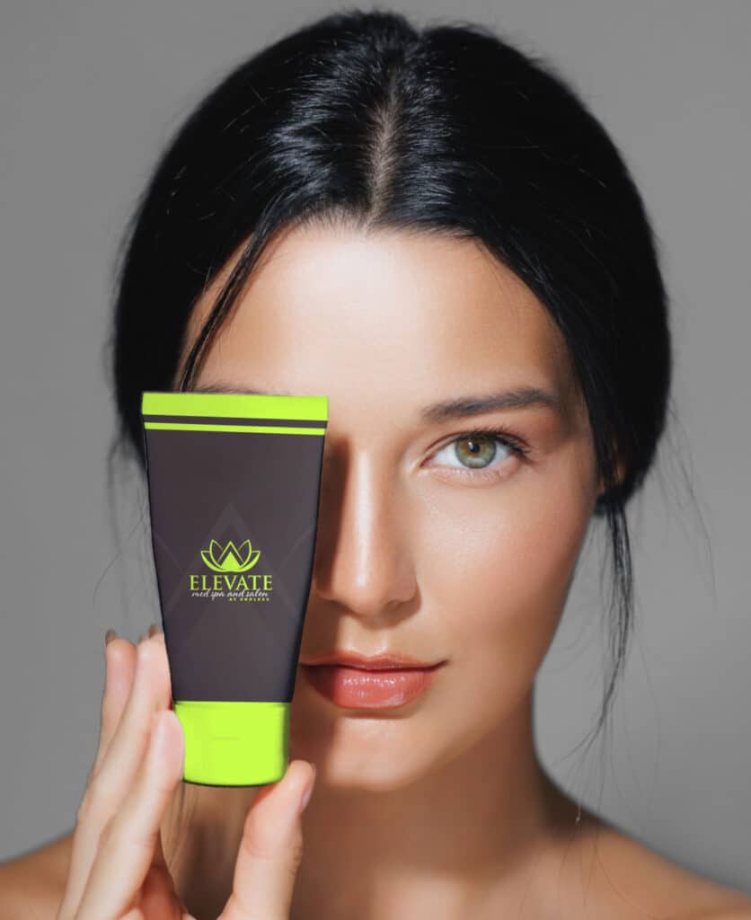 A woman holding a tube of Elevate skincare product in front of her face. She has long dark hair and balanced makeup, with a neutral background emphasizing her features and the product.