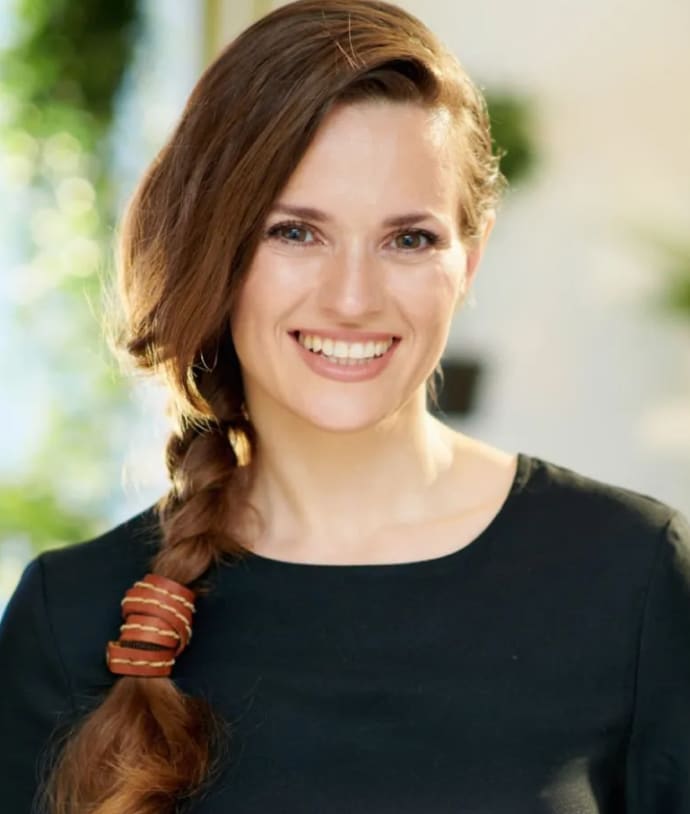 A woman with long brown hair styled in a loose braid, wearing a black top, smiling at the camera. The background features soft greenery and natural light, creating a bright atmosphere.