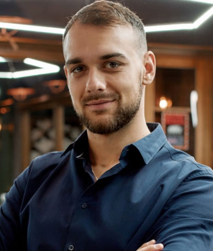 A man smiling confidently with his arms crossed, wearing a dark blue shirt. The background features warm lighting and a modern interior design, suggesting an upscale venue.
