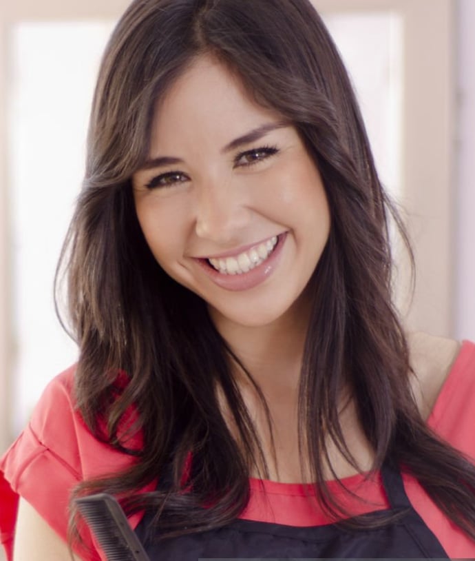 A woman with long dark hair smiles while wearing a red shirt and an apron. She holds a hair comb in one hand, standing in a bright, well-lit salon environment.
