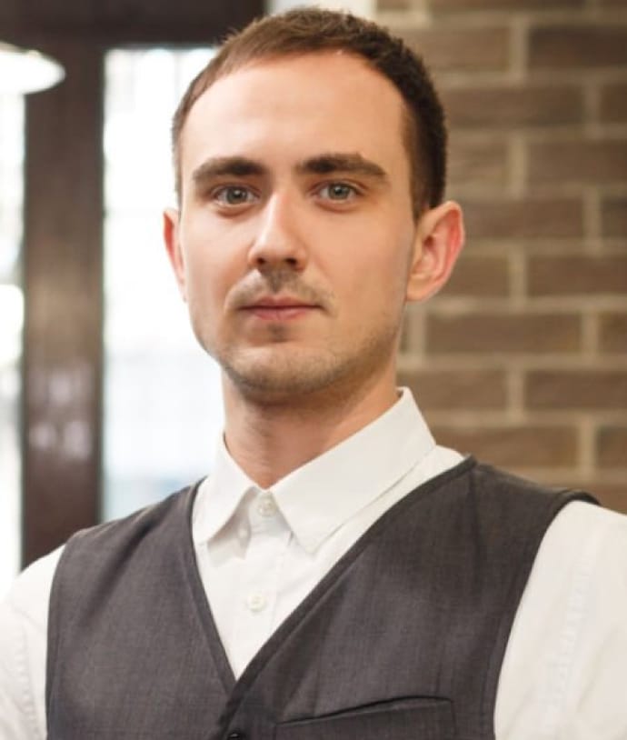 A man with short hair and a neutral expression, wearing a white shirt under a dark gray vest, stands in front of a brick wall background. The setting appears to be an indoor space with soft lighting.