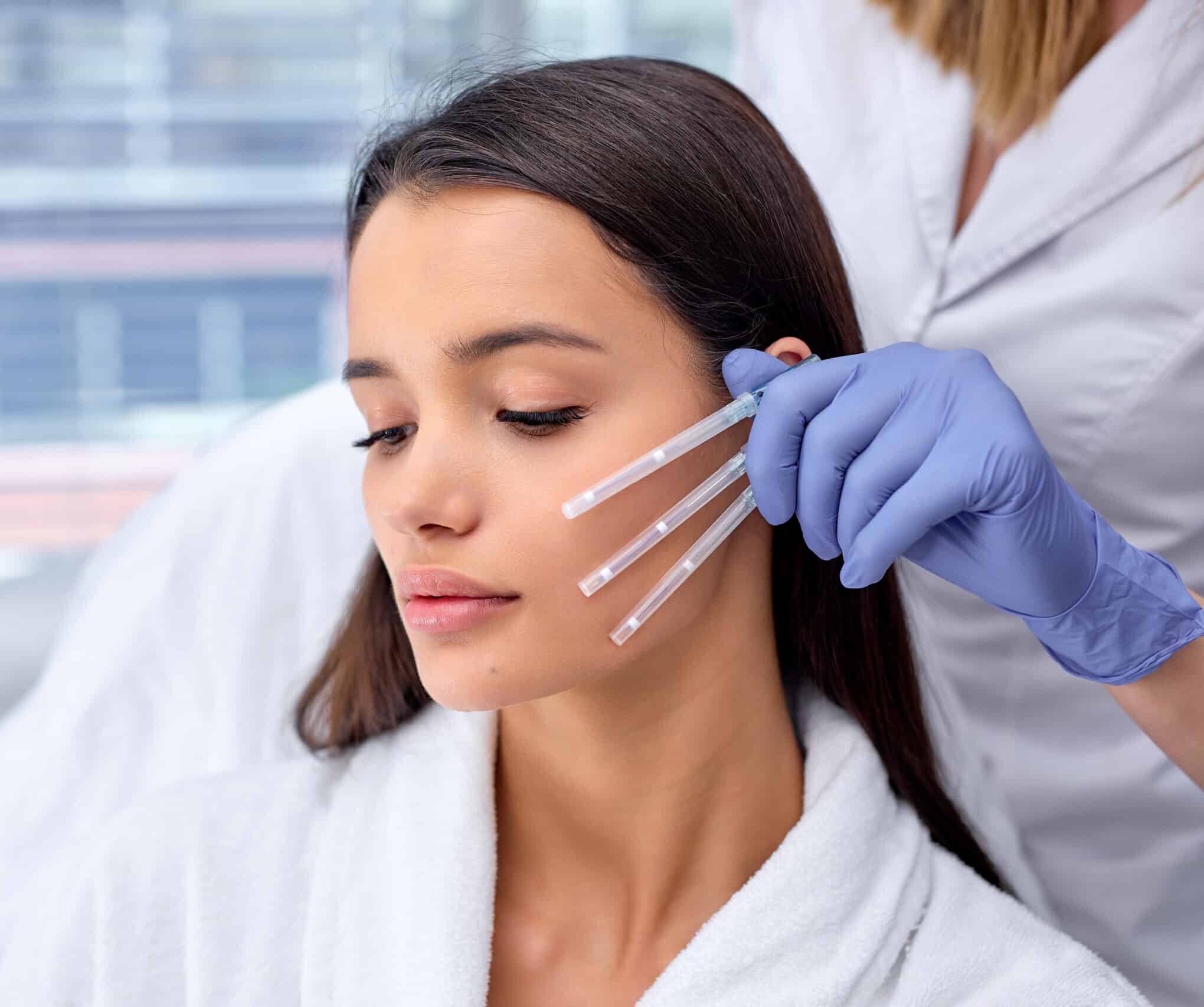 A woman in a white robe sitting in a treatment room, showing a calm expression. A professional in blue gloves is holding three clear applicators near her face, suggesting a cosmetic or skincare procedure.