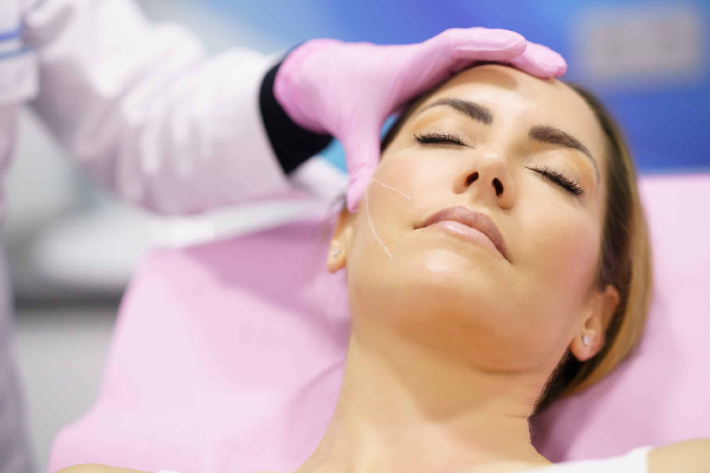 A woman resting on a treatment bed with closed eyes, receiving a cosmetic procedure. A clinician in a white coat and pink gloves is gently touching her face as part of the process in a clinical setting.