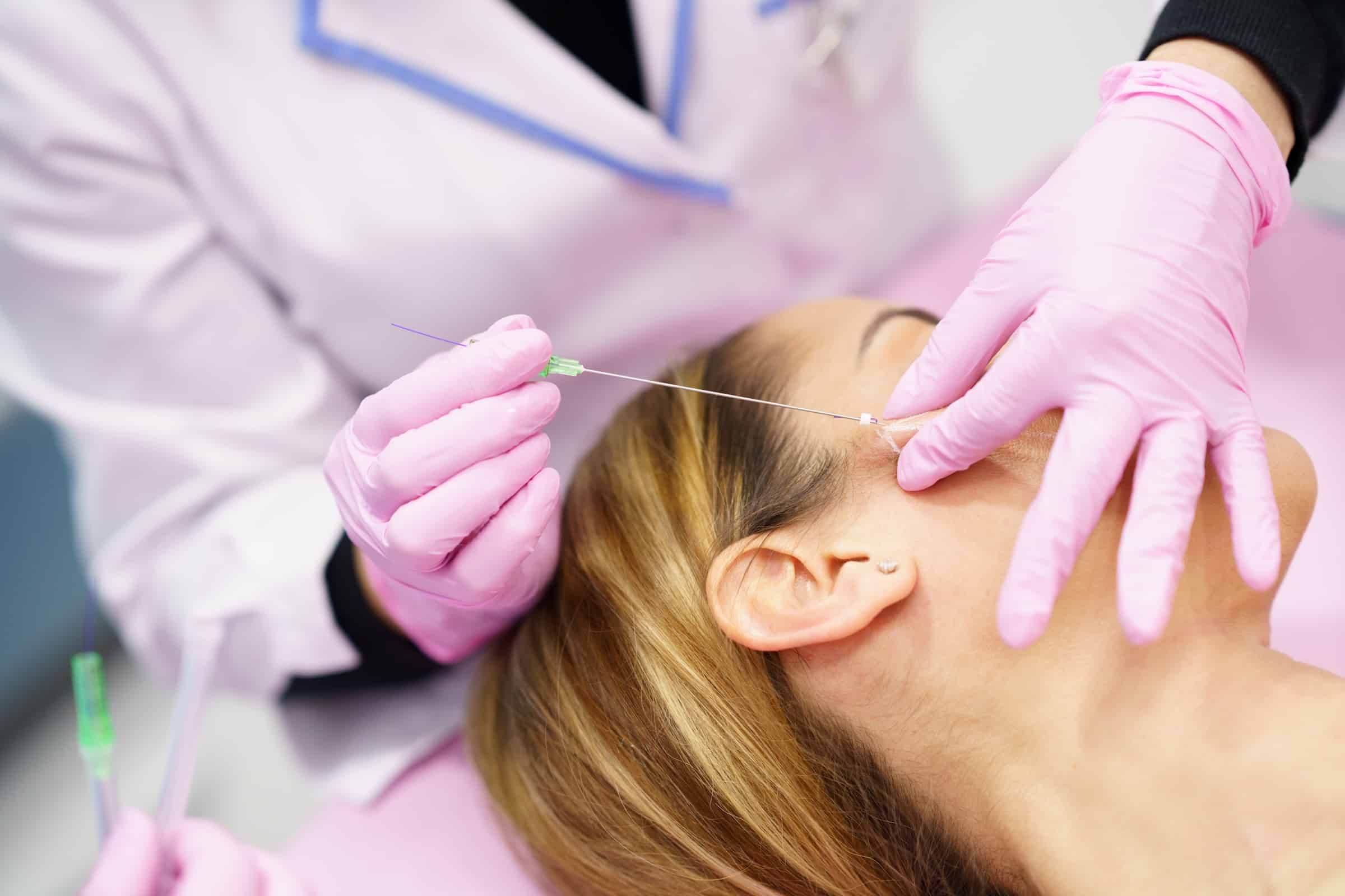 A cosmetologist wearing pink gloves performs a facial treatment on a patient lying down, focusing on the area around the patient's eye. The setting appears to be a clinical or spa environment.