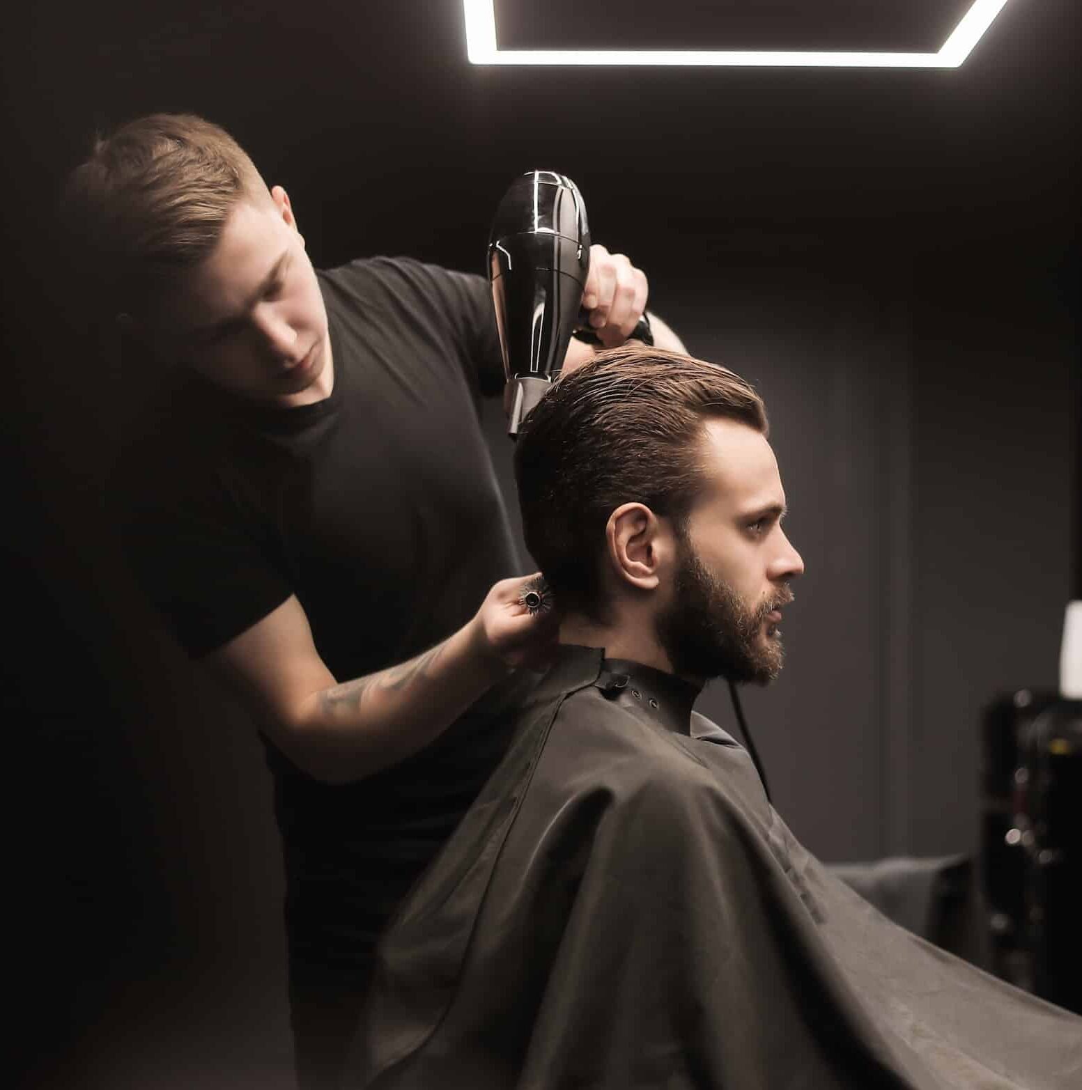 A hairstylist using a hairdryer on a client's head in a dimly lit salon. The client, with a beard and slicked-back hair, wears a black cape, while the stylist, dressed in a black shirt, focuses on the haircut.