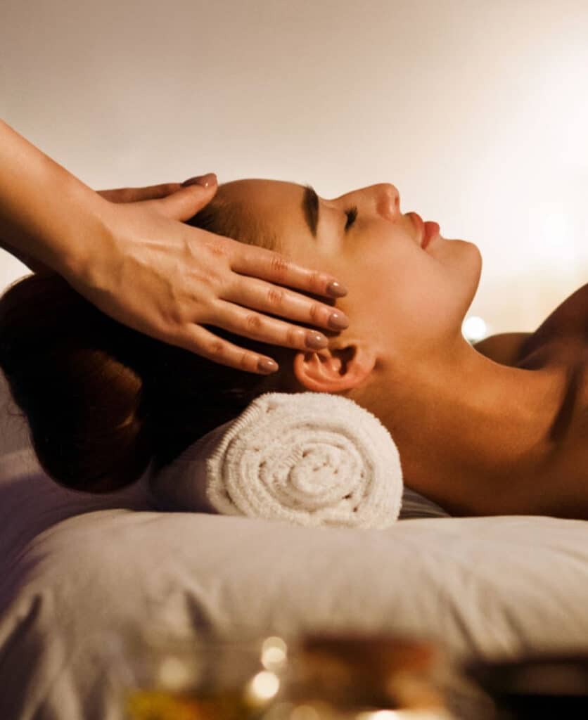 A woman receiving a head massage while lying on a massage table, with her head resting on a rolled towel. A therapist's hand gently touches her forehead, and soft lighting creates a relaxing atmosphere.