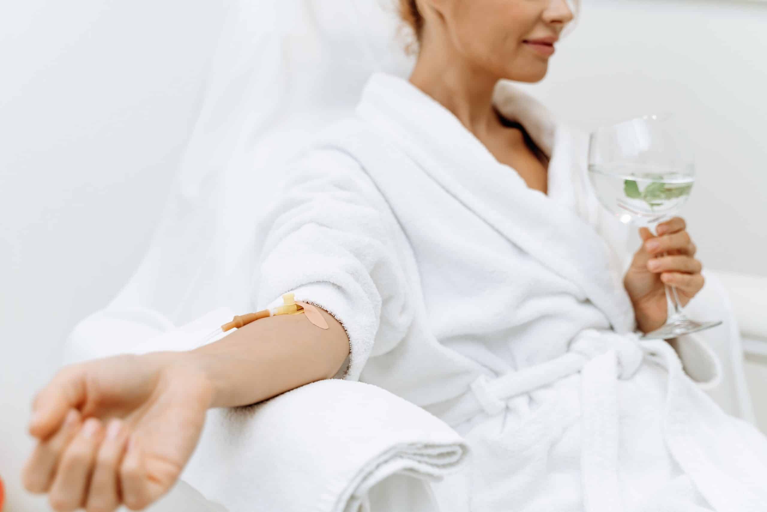 A woman in a white bathrobe sitting in a reclining chair, holding a glass of a green beverage with mint. An IV drip is attached to her arm, suggesting she is receiving treatment in a spa or wellness center.
