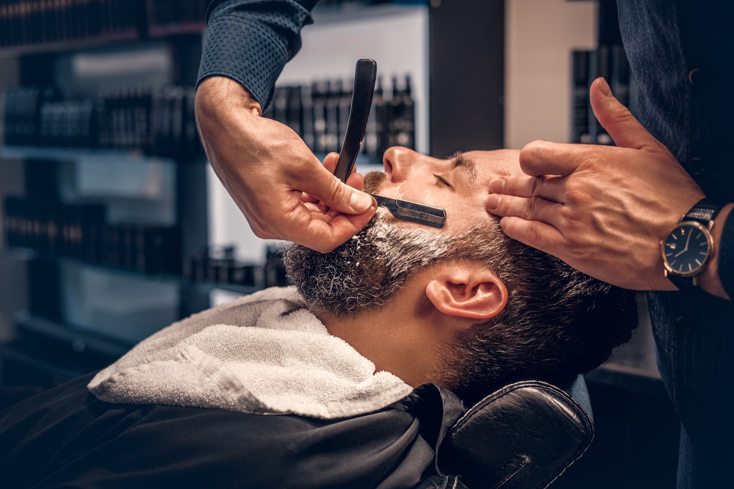 A barber using a straight razor to shave the beard of a man reclining in a salon chair, with products displayed on shelves in the background.