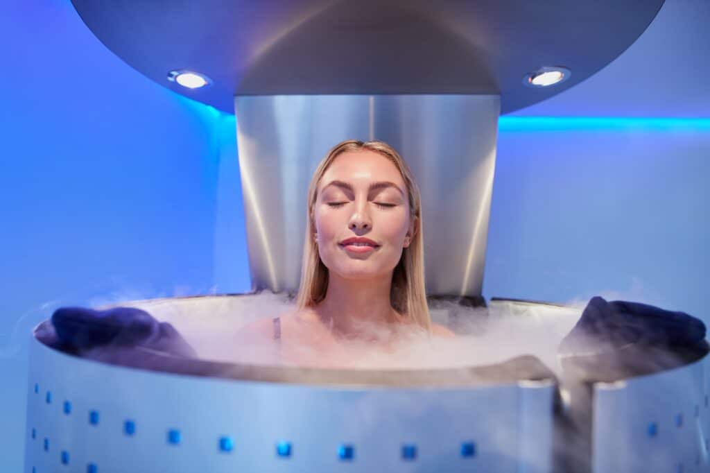 A woman with blonde hair relaxing in a cryotherapy chamber, surrounded by fog. The chamber features sleek metallic design and is illuminated by blue lights.