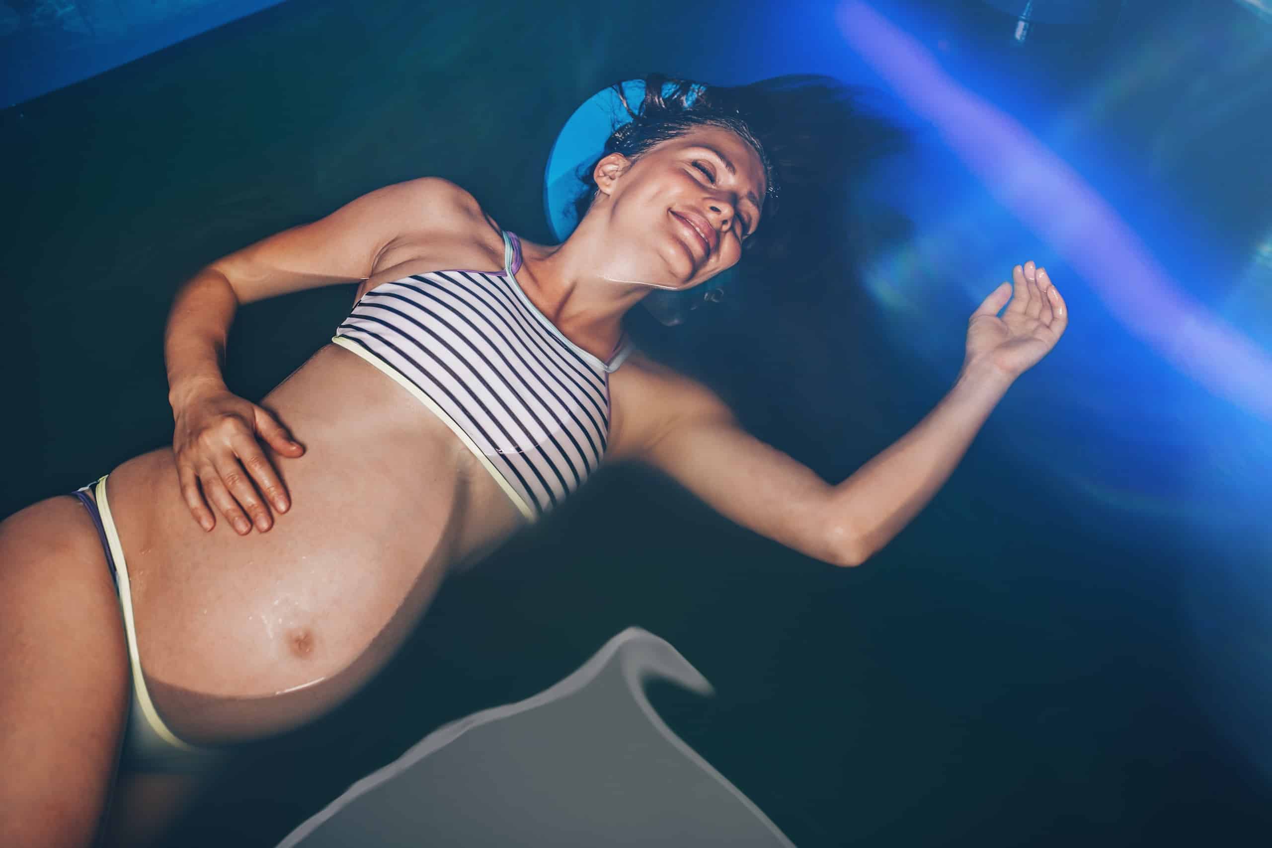 A pregnant woman in a striped bikini floating in water, with her hair spread out around her. She appears relaxed, facing upwards as light reflects on the water's surface.