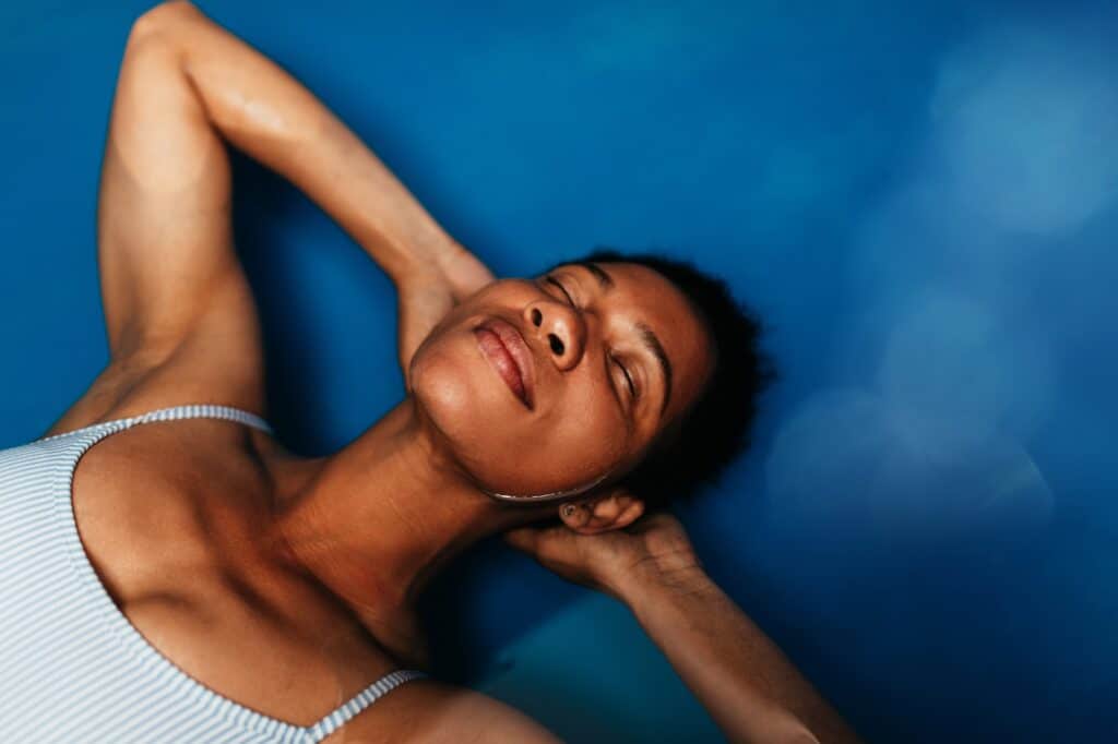 A woman lying back with her eyes closed, resting her hands behind her head. She has short dark hair and is wearing a light-colored swimsuit. The background is a smooth blue surface with soft lighting.
