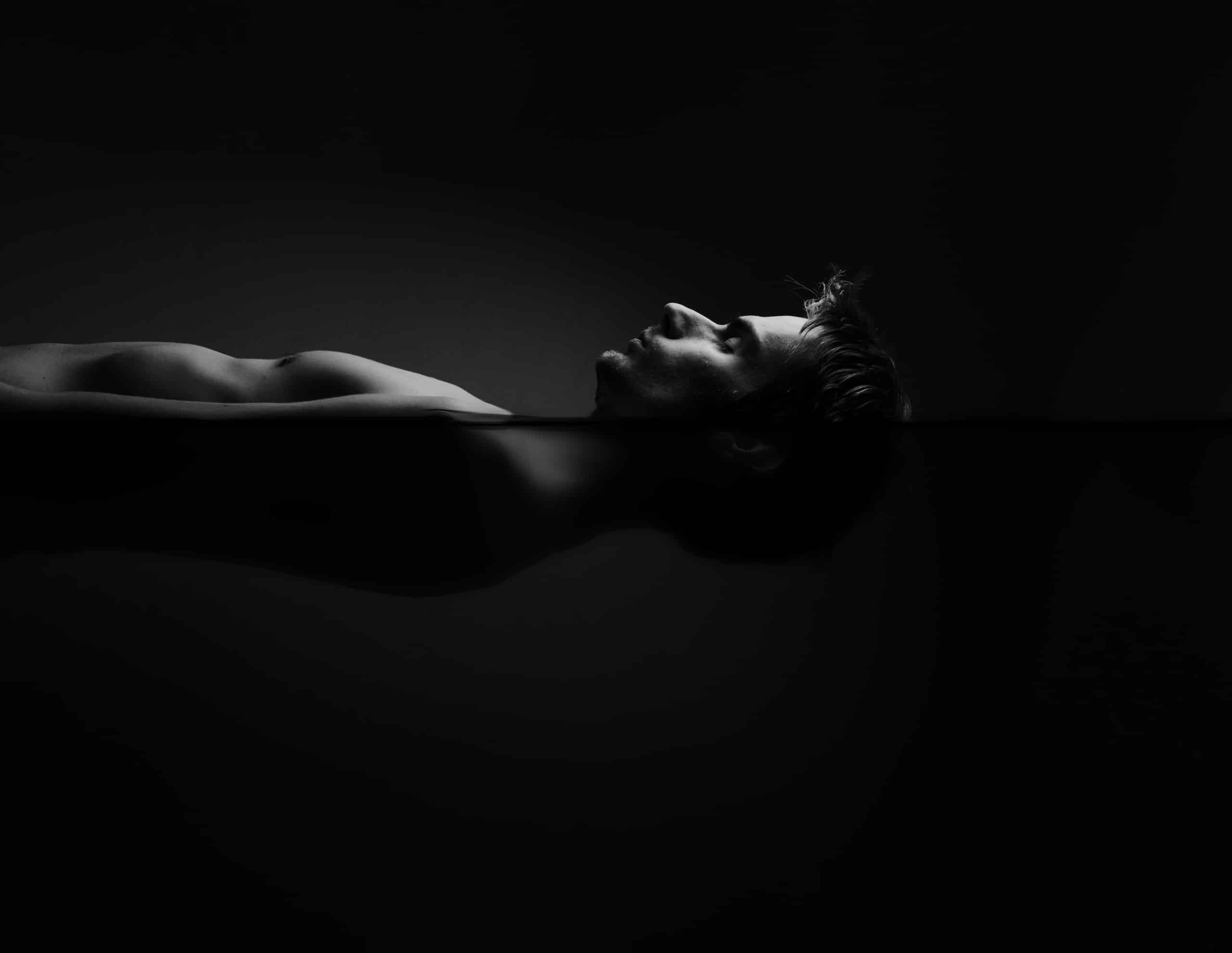A monochromatic image of a man floating in dark water. His body is partially submerged, with only his upper torso visible. The scene is illuminated softly, highlighting the contours of his face and body against the dark background.