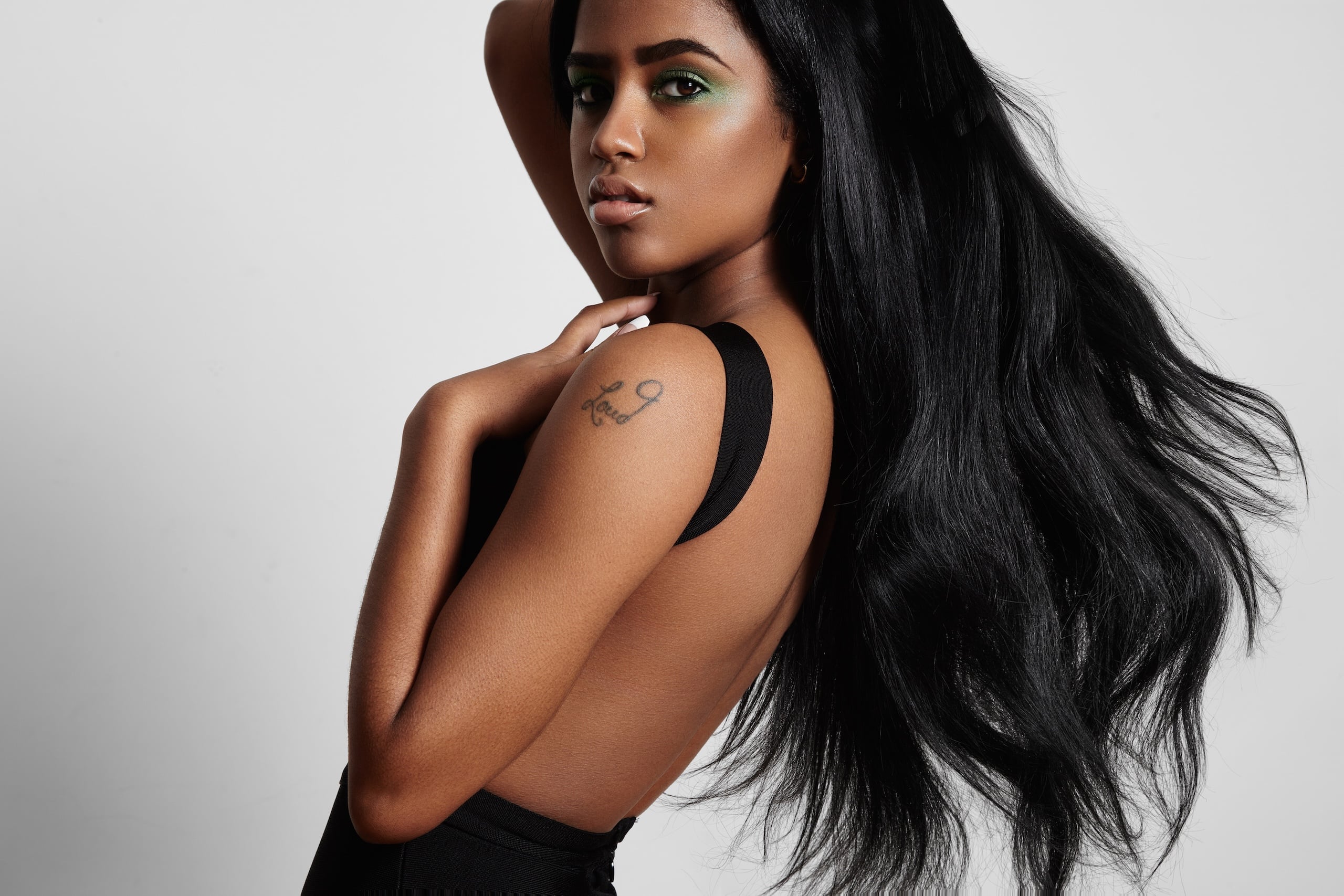 A woman with long black hair and green eye makeup poses with an expression of confidence. She has a tattoo on her shoulder and wears a black outfit, showcasing a stylish and bold look against a neutral background.
