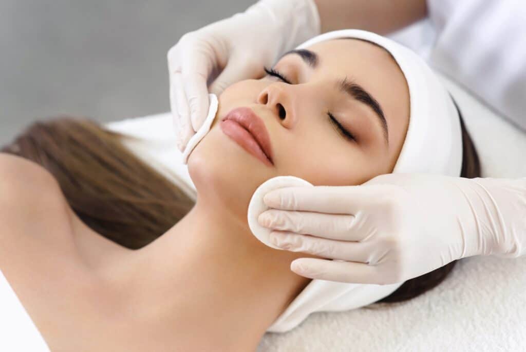 A woman receiving a facial treatment at a spa. A skincare professional, wearing gloves, gently applies cotton pads to her face, while the woman relaxes with her eyes closed. The setting features a soft, neutral background.