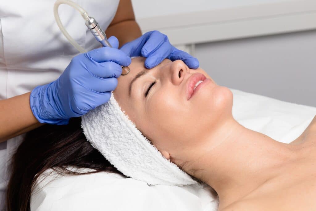 A woman receiving a facial treatment while lying on a bed. A professional, wearing blue gloves, uses a device on the woman's forehead. The woman has a towel around her head and appears relaxed.