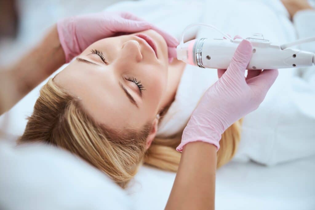A woman receiving a facial treatment at a spa, lying on a treatment table with her eyes closed. A professional in pink gloves is using a skin treatment device on her chin.
