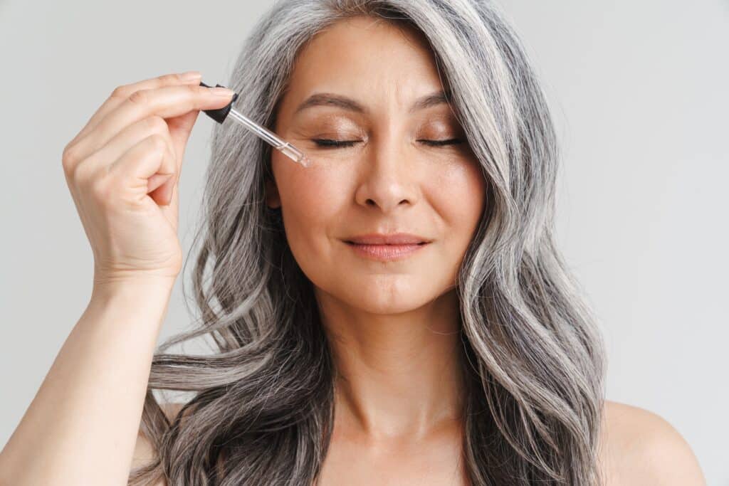 A woman with long gray hair applying a dropper of oil to her cheek while her eyes are closed, in a neutral-toned background.