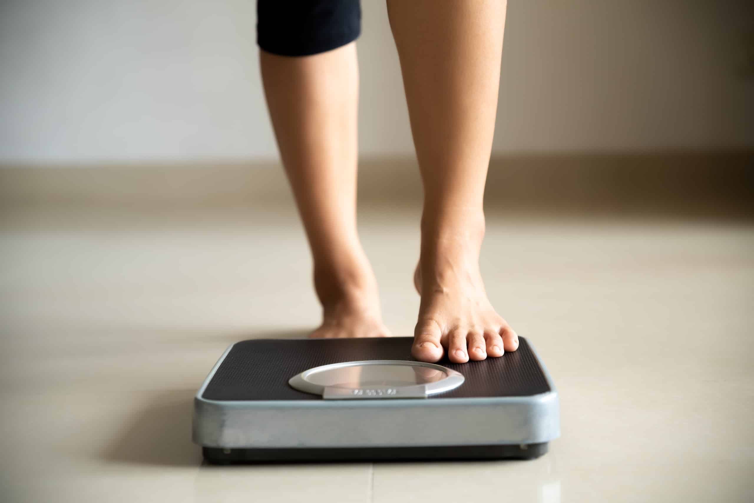 A person standing on a digital weighing scale with bare feet. The scale is on a smooth, light-colored floor, and the person's legs are partially visible, with black pants. The setting appears to be indoors.