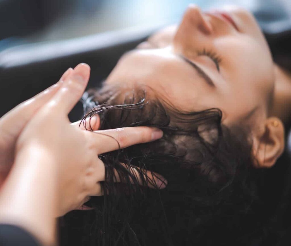 A close-up of a woman receiving a hair treatment, with a person's hand gently massaging her scalp. The woman appears relaxed, with her eyes closed. The background is softly blurred, emphasizing the intimate salon setting.