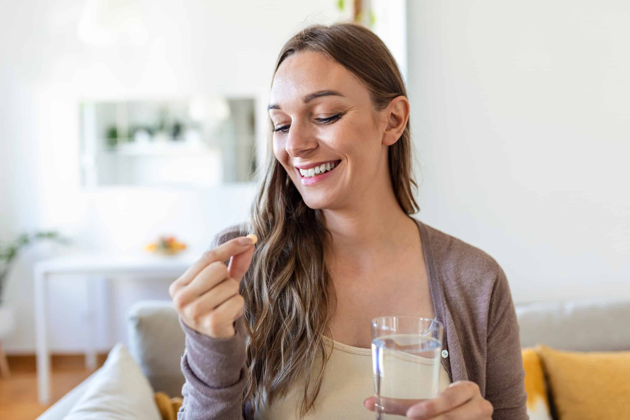 A woman sitting on a couch holds a clear glass of water in her right hand and examines a small tablet in her left hand. She has long brown hair and a slight smile, with a softly decorated living space in the background.