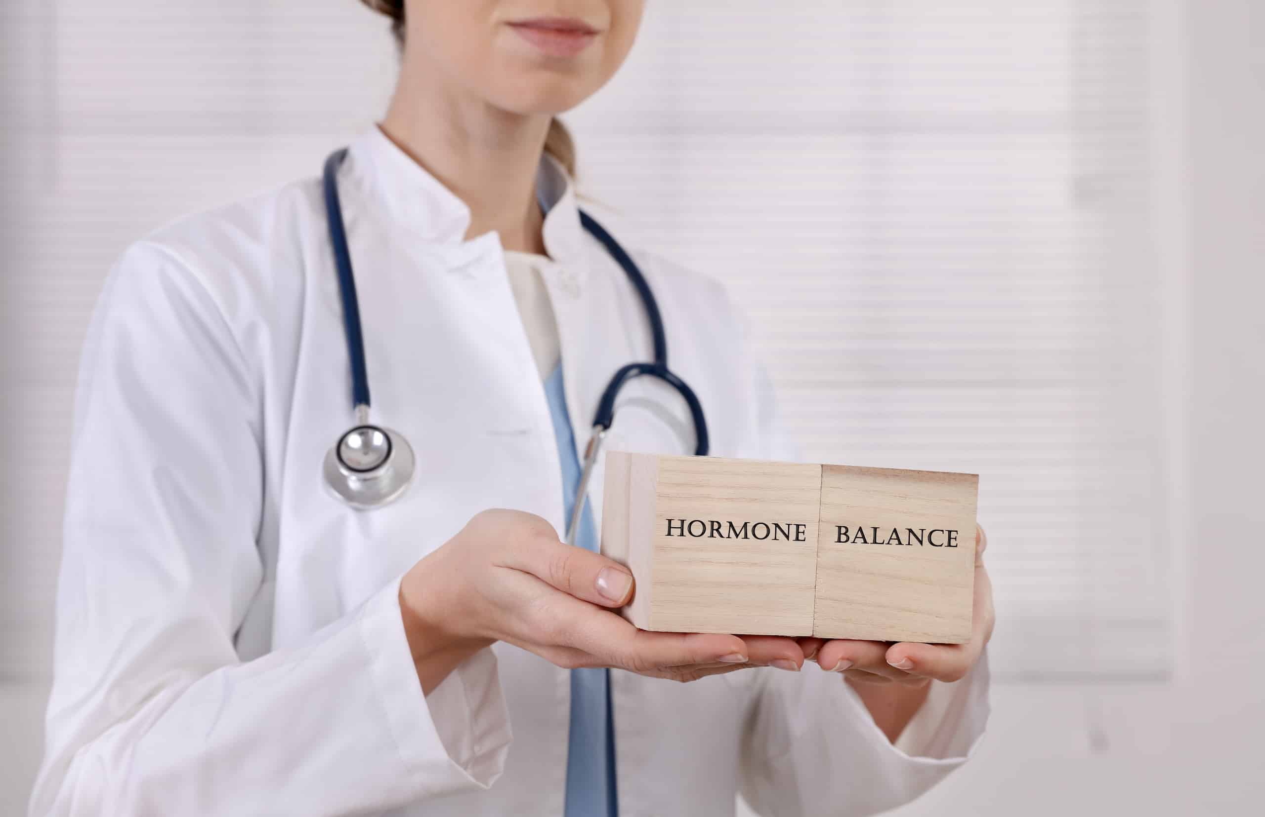 A healthcare professional in a white coat holds wooden blocks labeled "HORMONE" and "BALANCE". The background shows a blurred environment, emphasizing the focus on the text on the blocks.