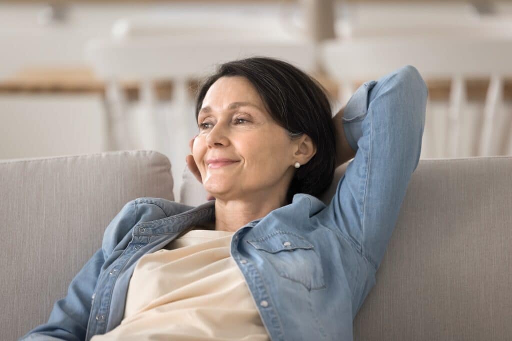 A middle-aged woman lounging on a light-colored sofa, wearing a denim jacket and a light top, with her hands behind her head and a calm expression. The background features a bright and airy living space.