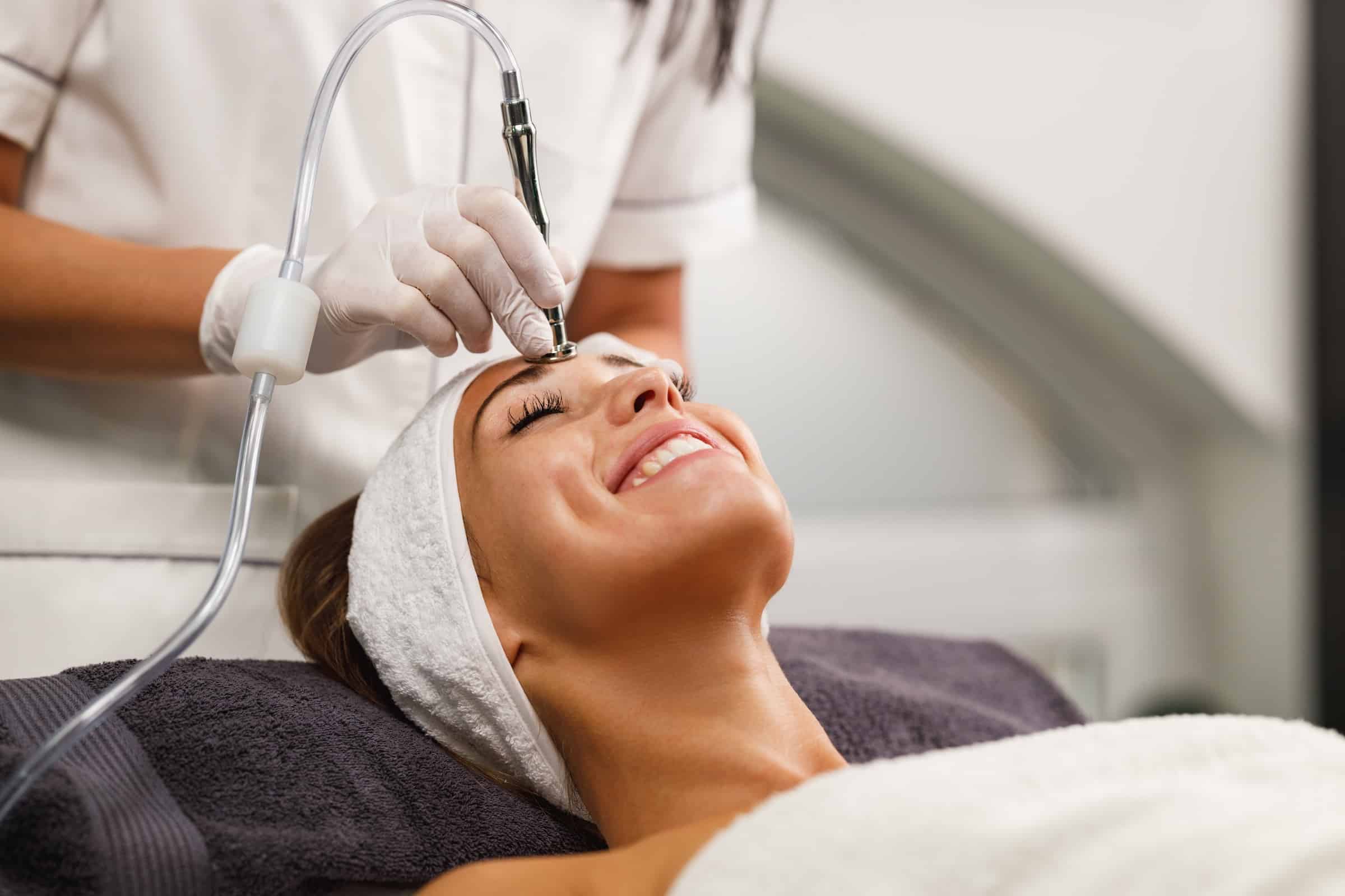 A woman receiving a facial treatment at a spa, smiling as a technician operates a device on her forehead. She is lying on a treatment bed, covered with a towel, with a headband to keep her hair away from her face.