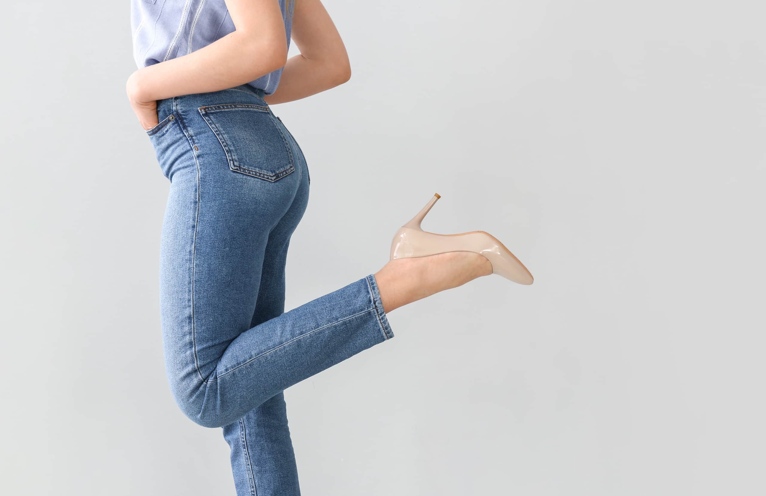 A person wearing light blue high-waisted jeans and a gray top is posing with one leg raised, showcasing beige high-heeled shoes. The background is plain and light, emphasizing the outfit.
