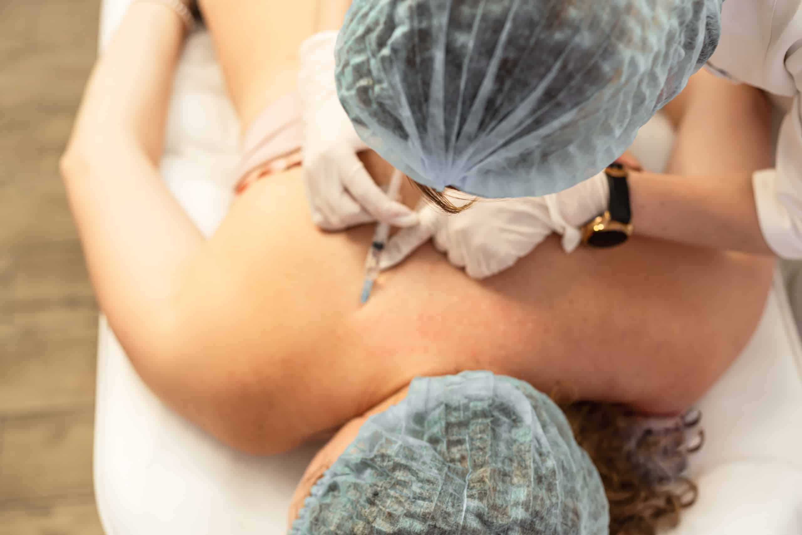 A medical professional in gloves and a cap injects a substance into the shoulder of a patient lying on a treatment table. The patient is positioned face down, with only their back visible, while the healthcare worker focuses on the procedure.