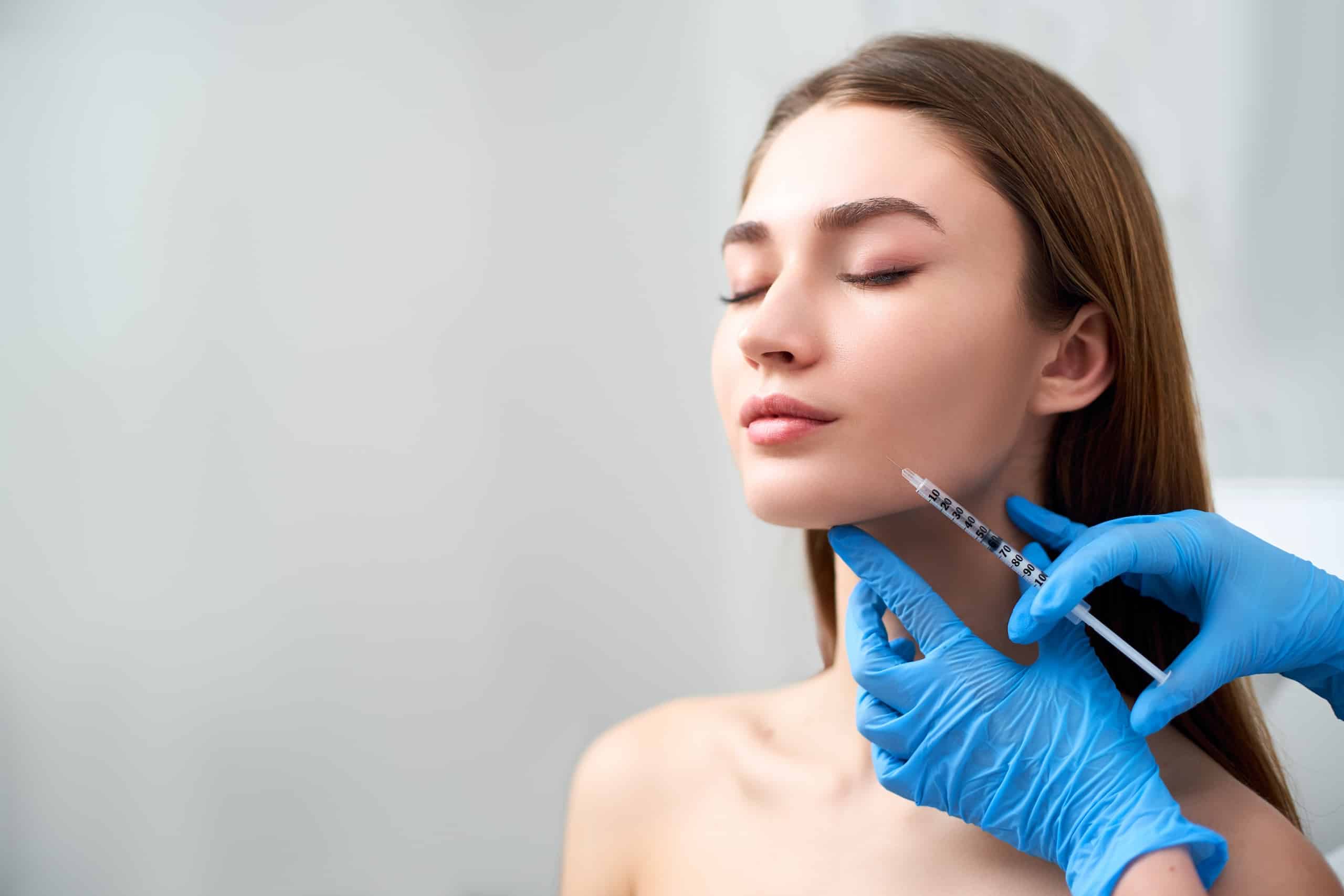 A young woman with closed eyes sitting calmly while a medical professional in blue gloves administers an injectable treatment to her jawline. The setting appears to be a clinical environment, with neutral tones and soft lighting.