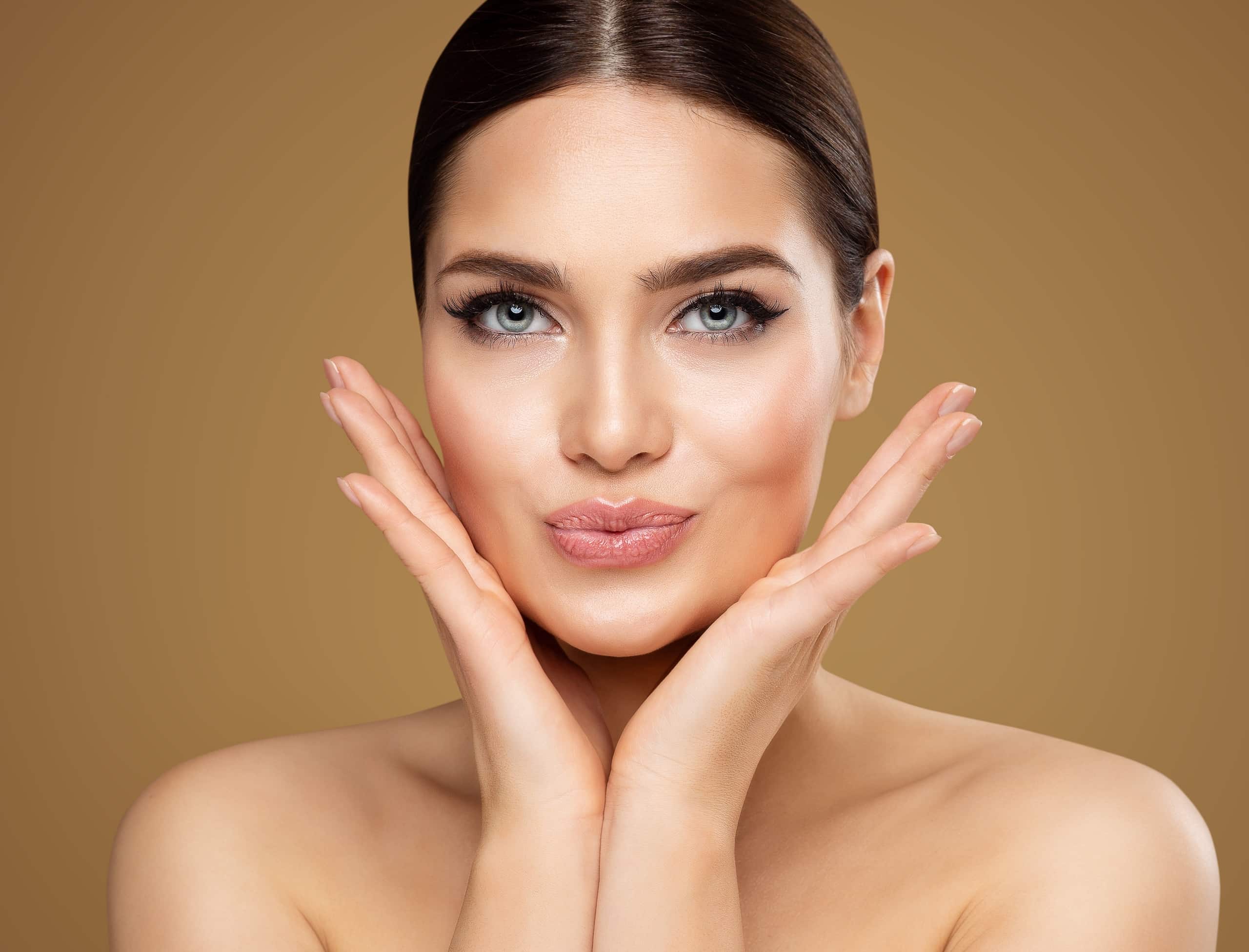 A woman with medium skin tone and straight dark hair poses against a gold background. She is holding her face with both hands, showcasing her clear skin and subtle makeup, featuring defined eyes and nude lips.