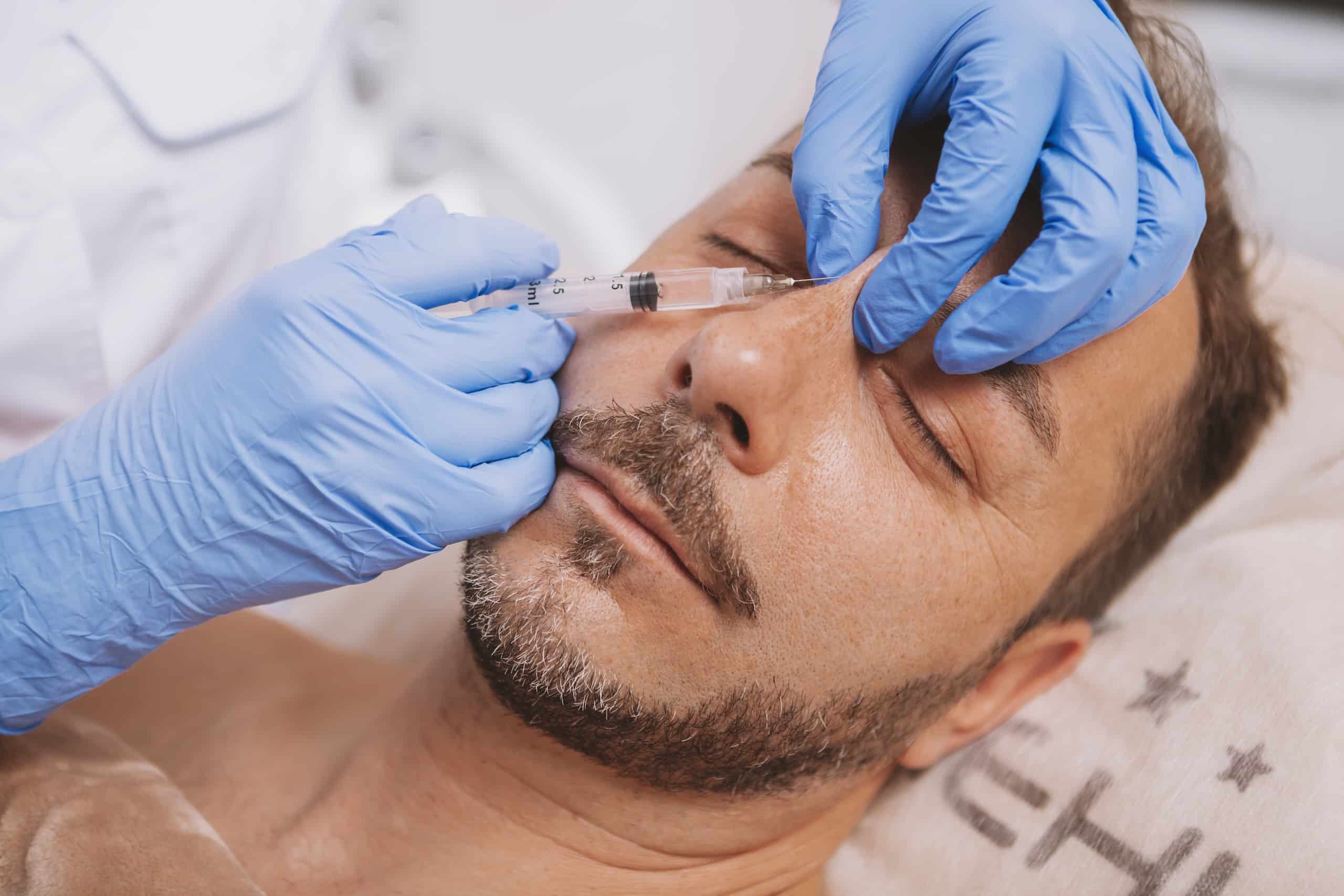 A close-up of a man receiving an injection on the side of his face, administered by a practitioner wearing blue gloves. The man has a beard and is lying on a treatment table with a towel draped over his torso.