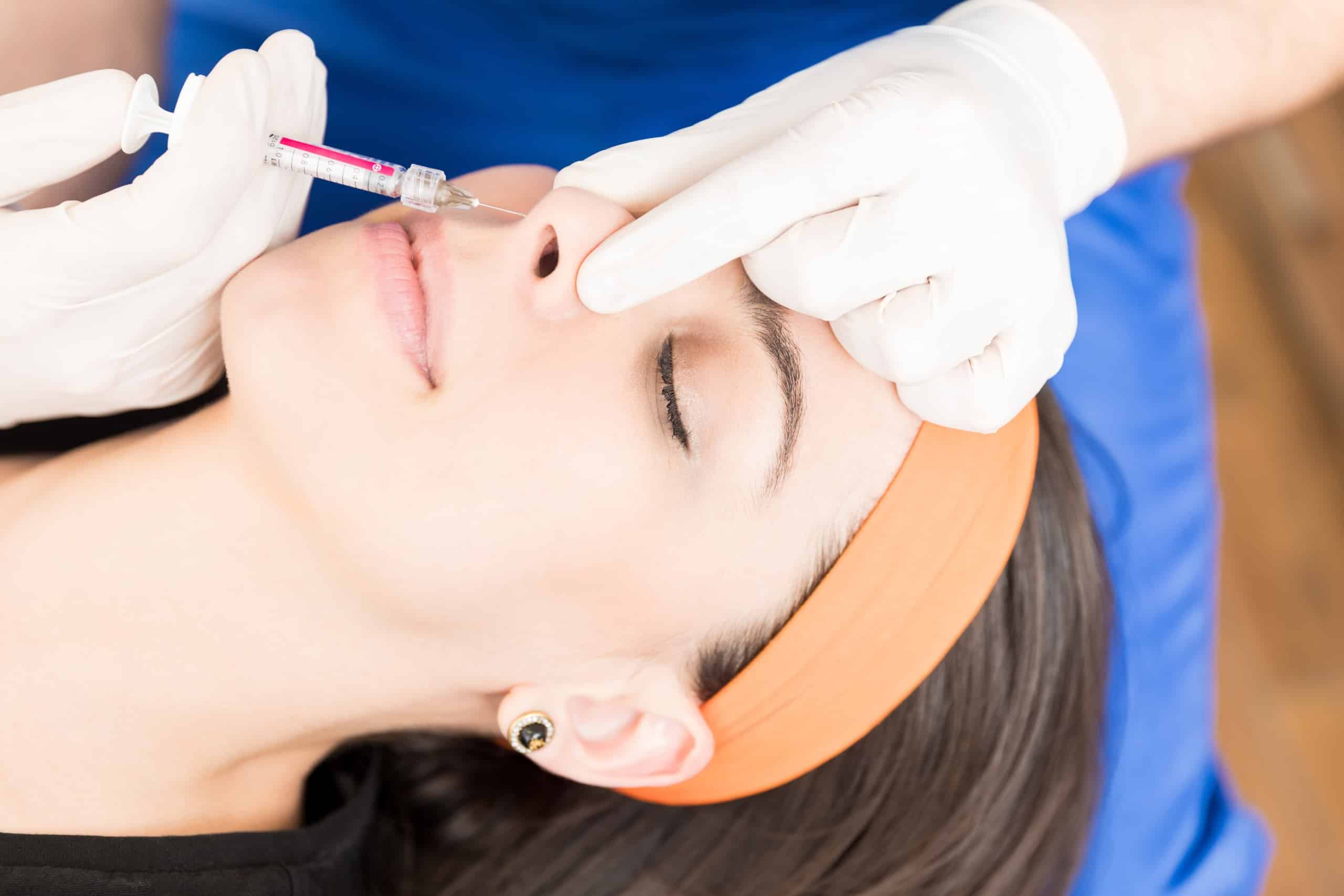 A woman with long dark hair and an orange headband is lying down with her eyes closed. A healthcare professional in white gloves administers an injection near her nose using a syringe. The setting appears to be a clinical environment.