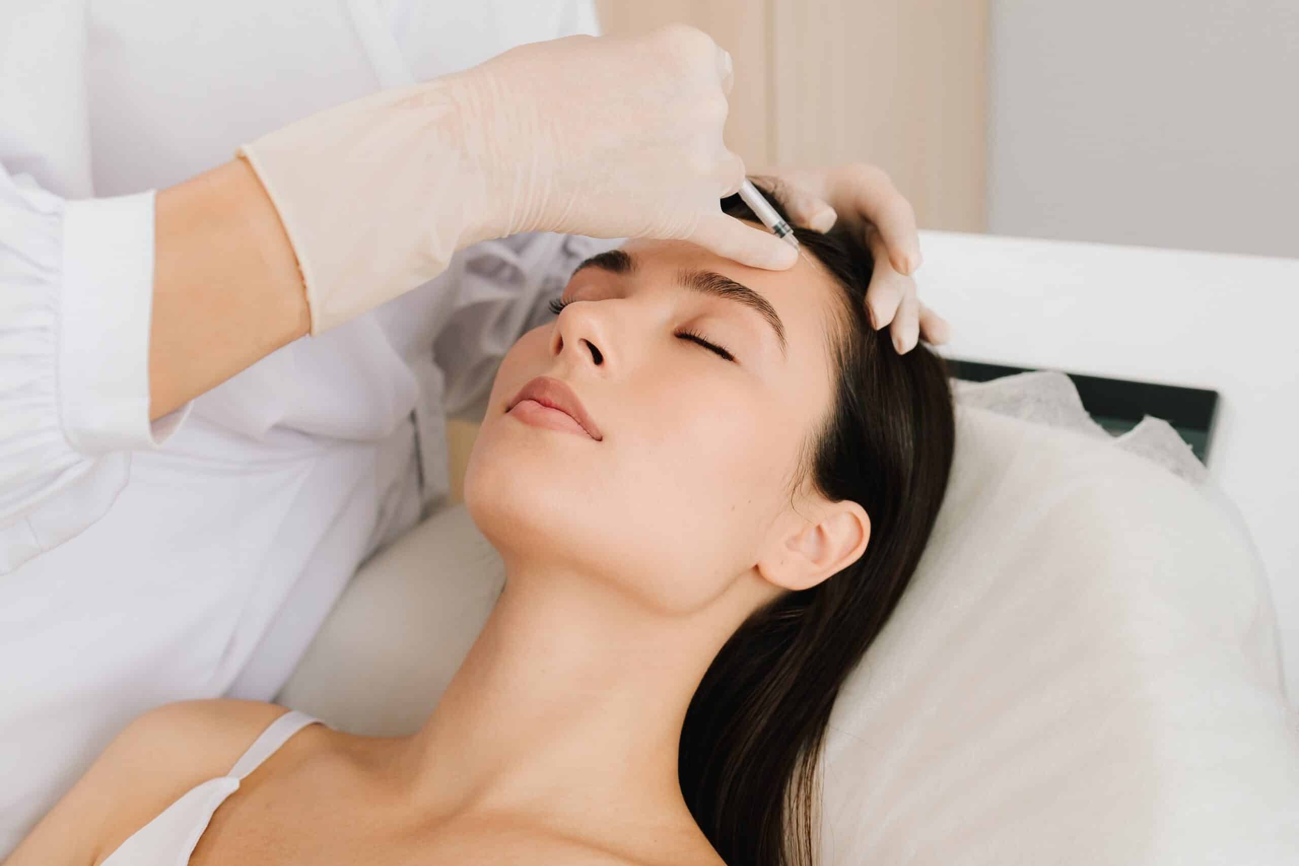 A woman receiving a cosmetic treatment in a clinic, with a gloved professional injecting a substance into her forehead. The patient has her eyes closed and is resting on a treatment bed.