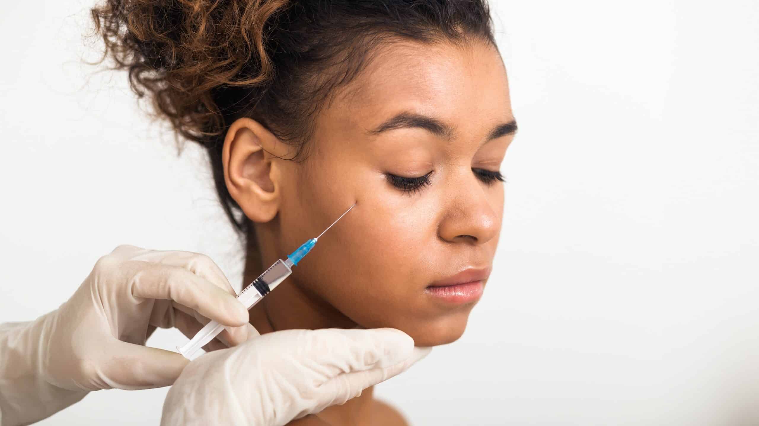 A close-up of a person receiving a cosmetic injection, with a gloved hand holding a syringe near their face. The individual has medium-length, curly hair and a neutral expression, against a plain background.