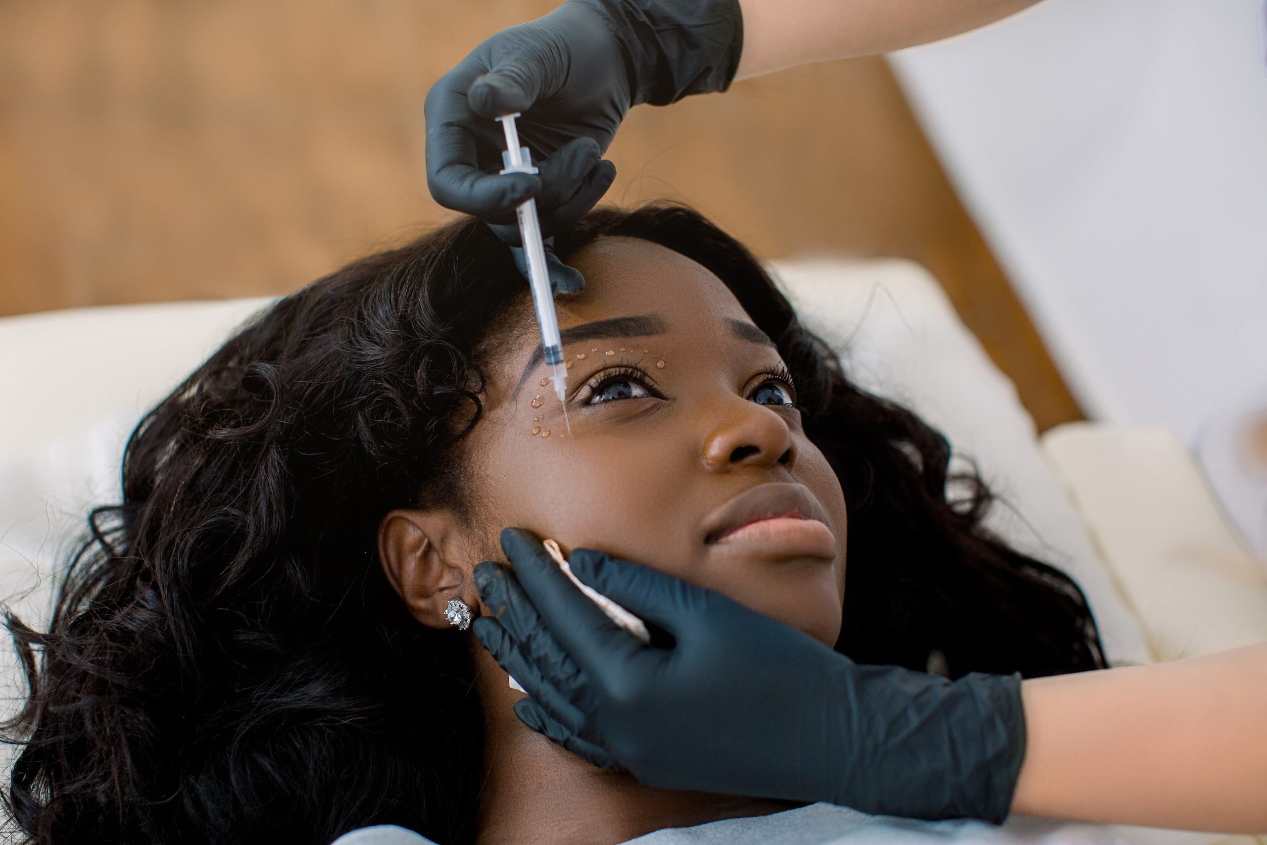 A woman lying on a treatment bed while a practitioner, wearing gloves, administers a cosmetic injection to her forehead. The setting appears to be a clean, medical or beauty treatment room.