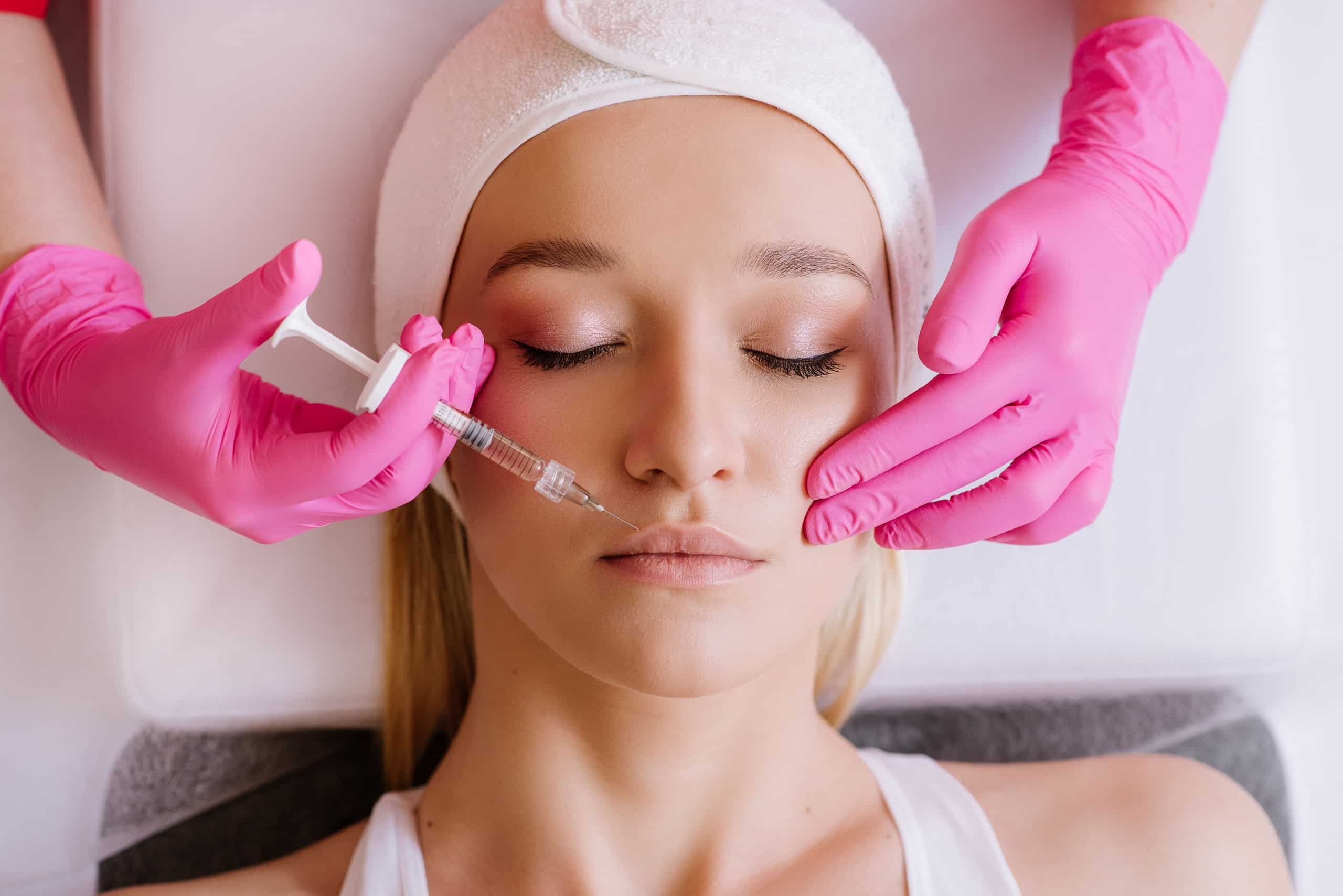 A woman receiving a cosmetic treatment in a spa-like setting. She is lying on a treatment table, eyes closed, as a practitioner in pink gloves administers an injection near her lips. The practitioner's hand is holding a syringe, and they are focused on the procedure.