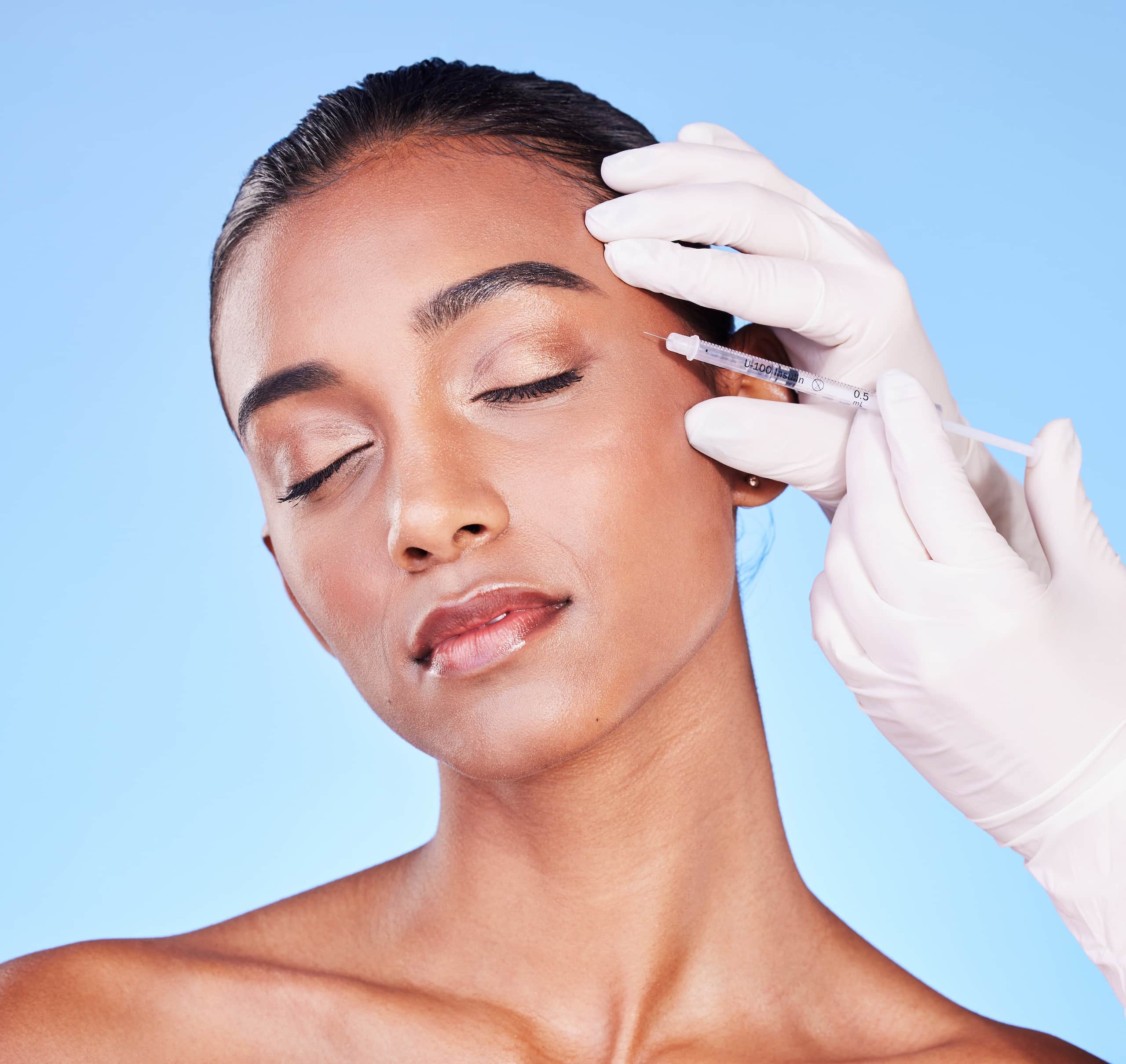 A woman with closed eyes is receiving an injection on her forehead from a healthcare professional wearing white gloves. The background is a light blue color, and the woman has a natural makeup look with a smooth complexion.