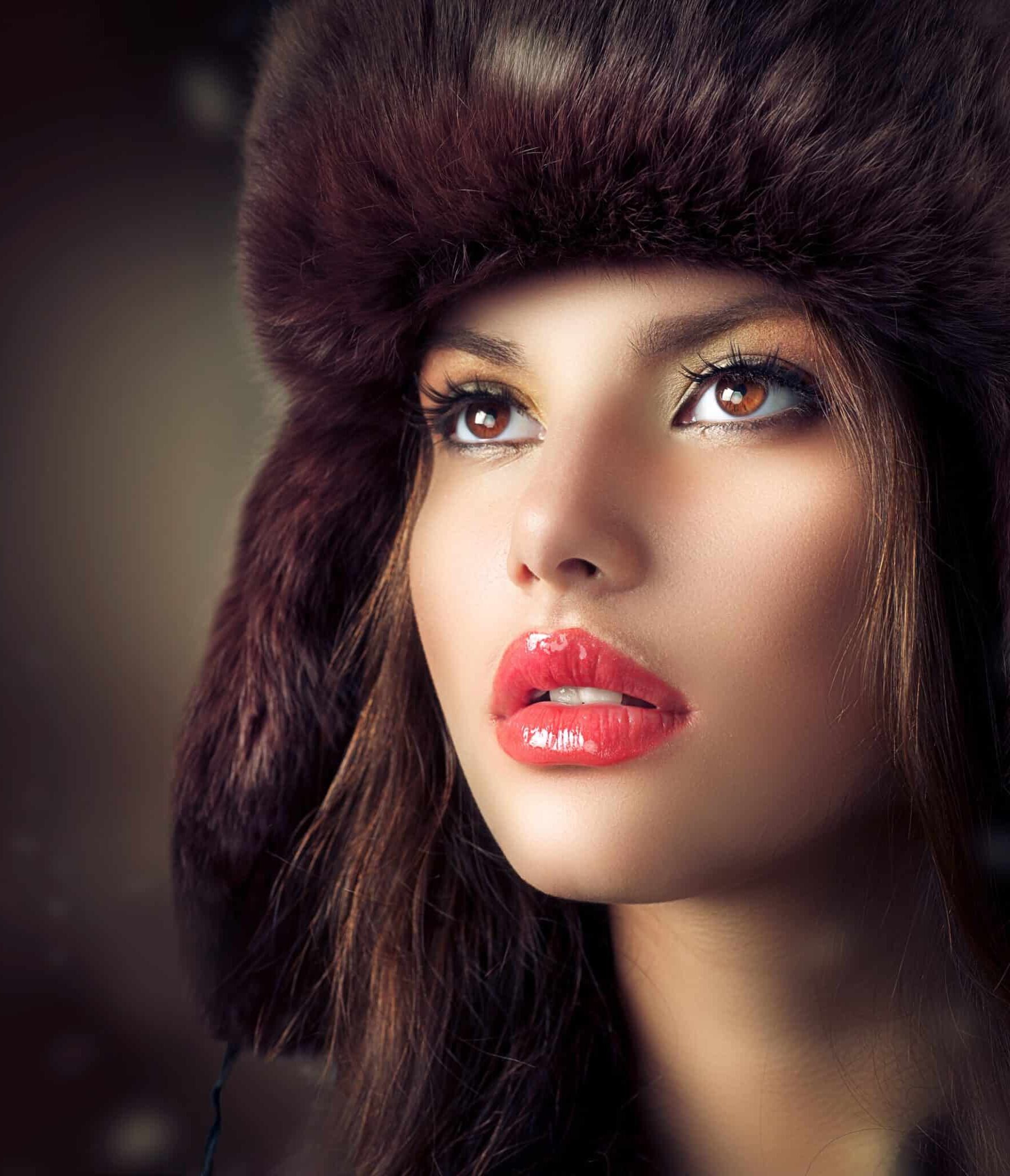 A close-up portrait of a woman with long dark hair wearing a dark fur hat. She has glossy red lips and striking brown eyes, looking upward with a neutral expression. The background is softly blurred, highlighting her features.