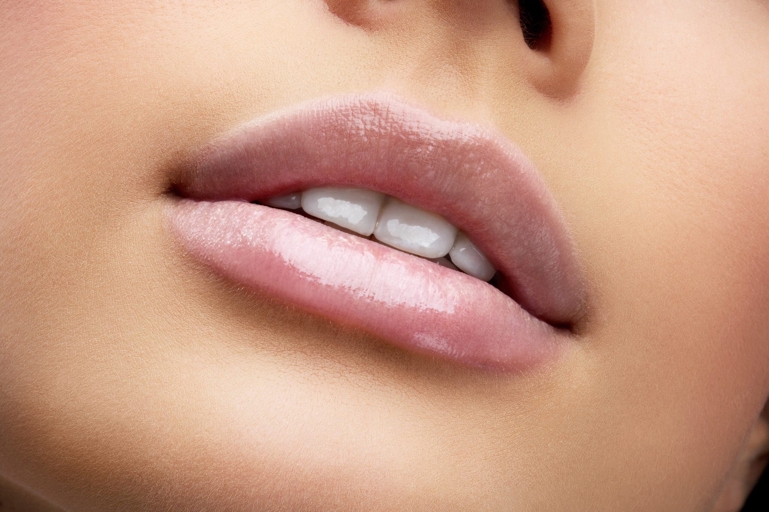Close-up view of soft, slightly full lips with a light pink gloss. The lower lip is fuller, and the teeth are visible. The background is blurred to emphasize the lips and skin tone, creating a smooth, clear appearance.
