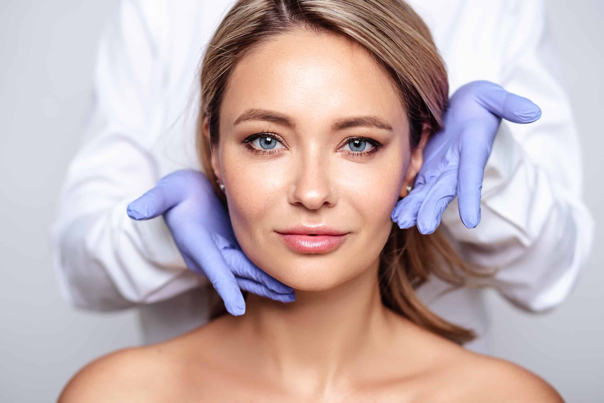A woman with shoulder-length blonde hair is sitting against a neutral background. A person wearing a white coat and blue gloves is gently placing their hands on the woman's face, indicating a cosmetic treatment or consultation.