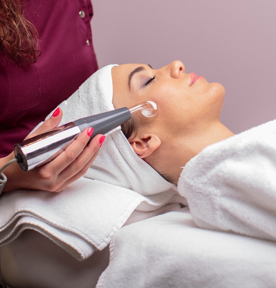 A woman receiving a facial treatment in a spa, lying on a treatment table with a towel draped over her shoulder. A therapist holds a small device near her face, applying a skin care treatment. The background is softly lit in neutral tones.