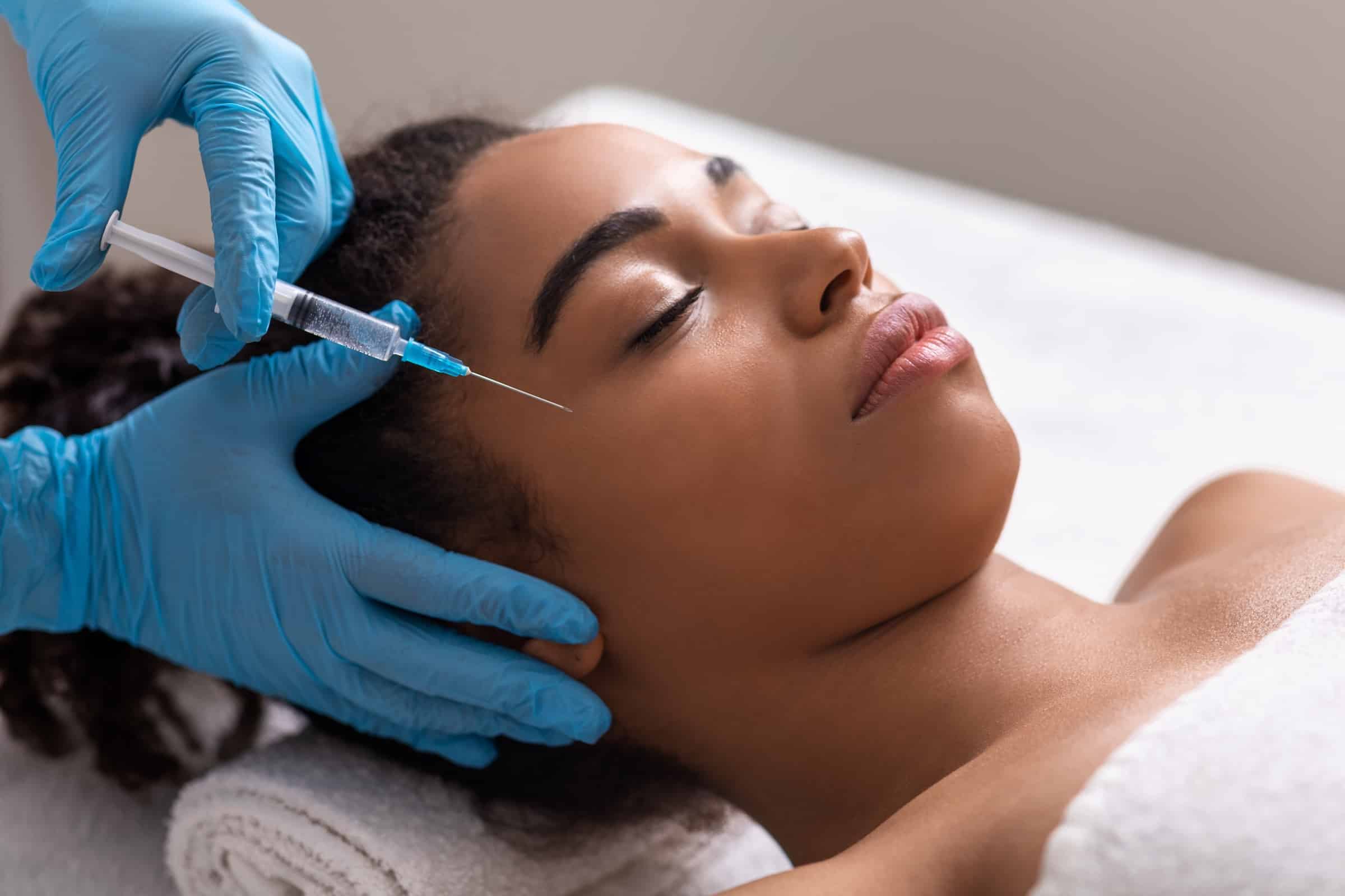 A healthcare professional in blue gloves administering an injection on the forehead of a relaxed woman lying on a treatment table, with her eyes closed and covered with a white towel.
