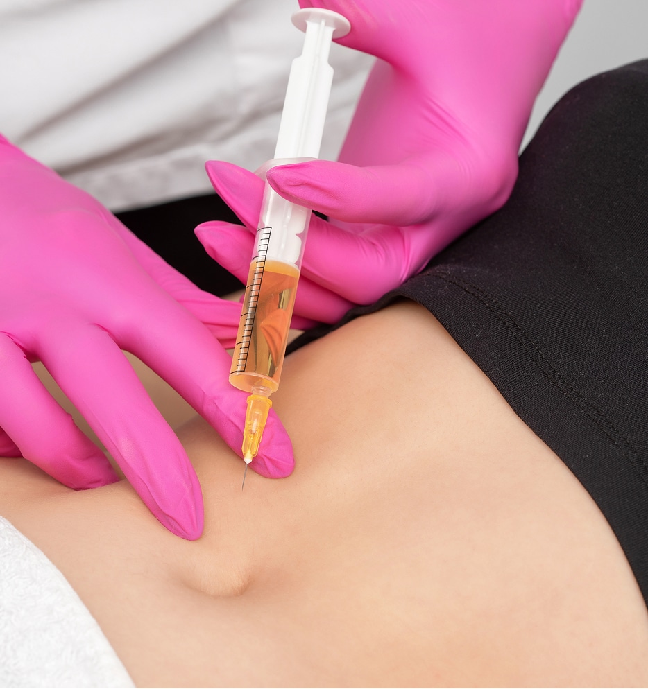 A close-up of a healthcare professional in pink gloves administering a syringe filled with a yellow liquid into the abdomen of a patient, who is lying down with a white cloth draped over part of their torso.
