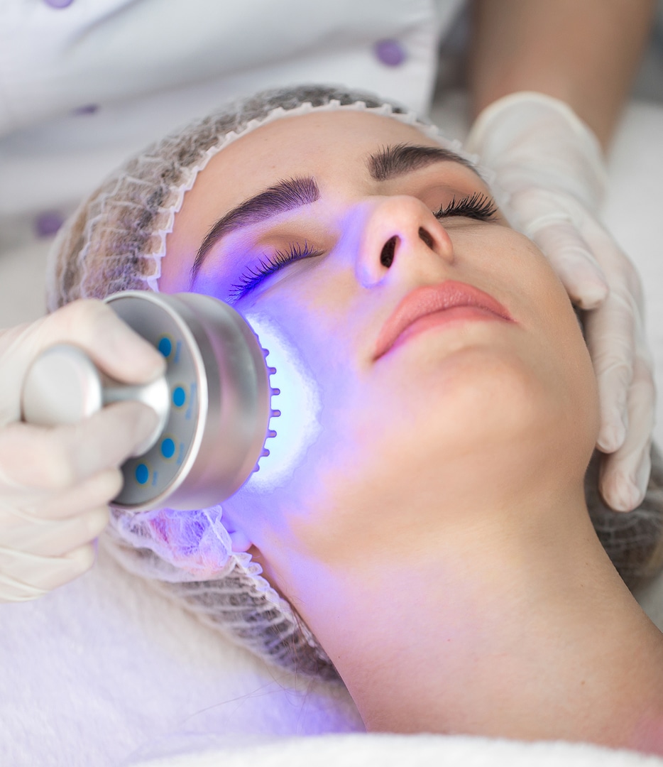 A woman receiving a facial treatment in a spa setting, with a professional using a blue light therapy device on her cheek. The woman is lying down with her eyes closed and wearing a hairnet.